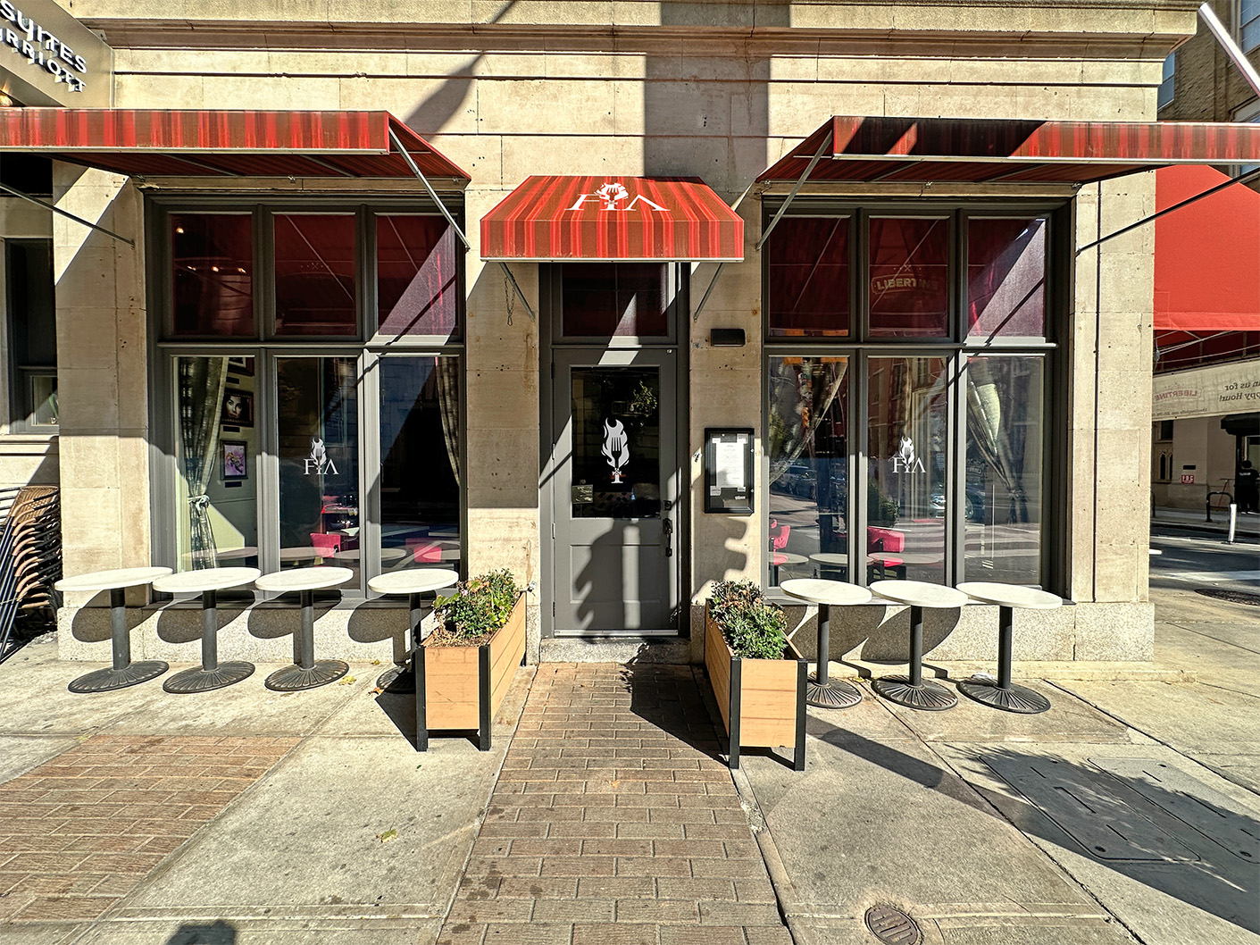 Exterior of FIA with red and orange striped awnings, outdoor round tables, and two wooden planter boxes flanking the entrance door.