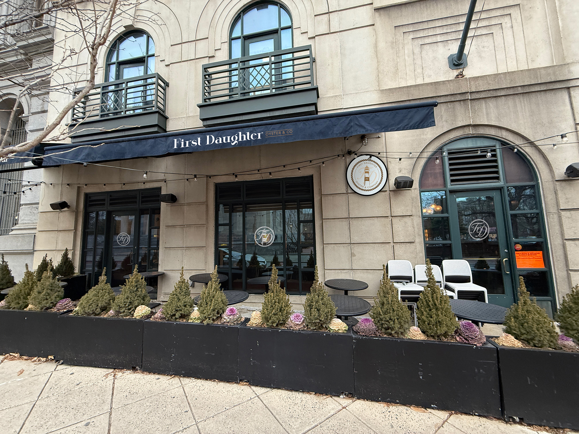 Outdoor seating area with tables, chairs, and decorative plants in front of a beige building with arched windows and a black awning that reads 'First Daughter Oyster & Co.'