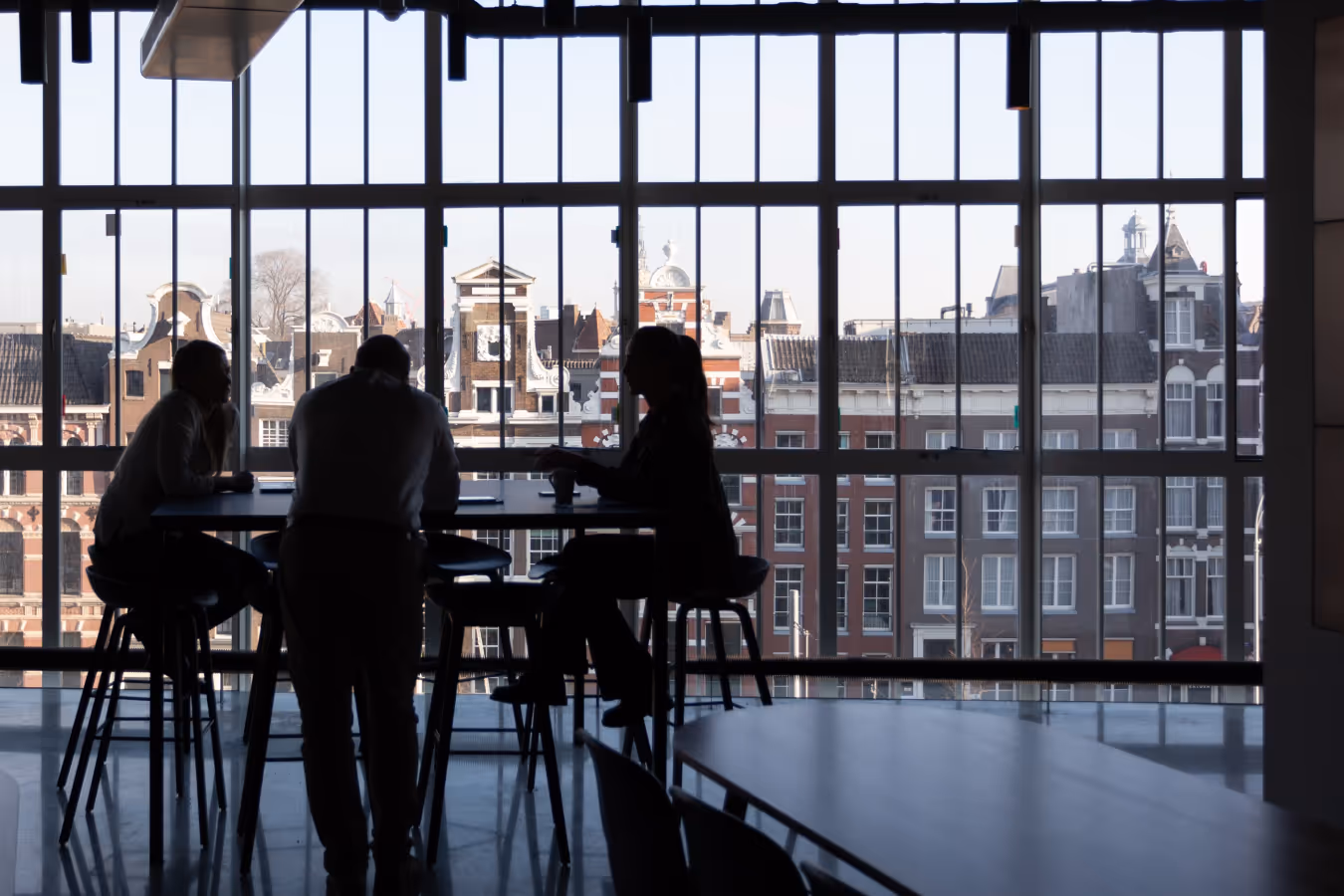 Silhouettes of three people sitting and standing around a high table by large windows with a view of historic European buildings.
