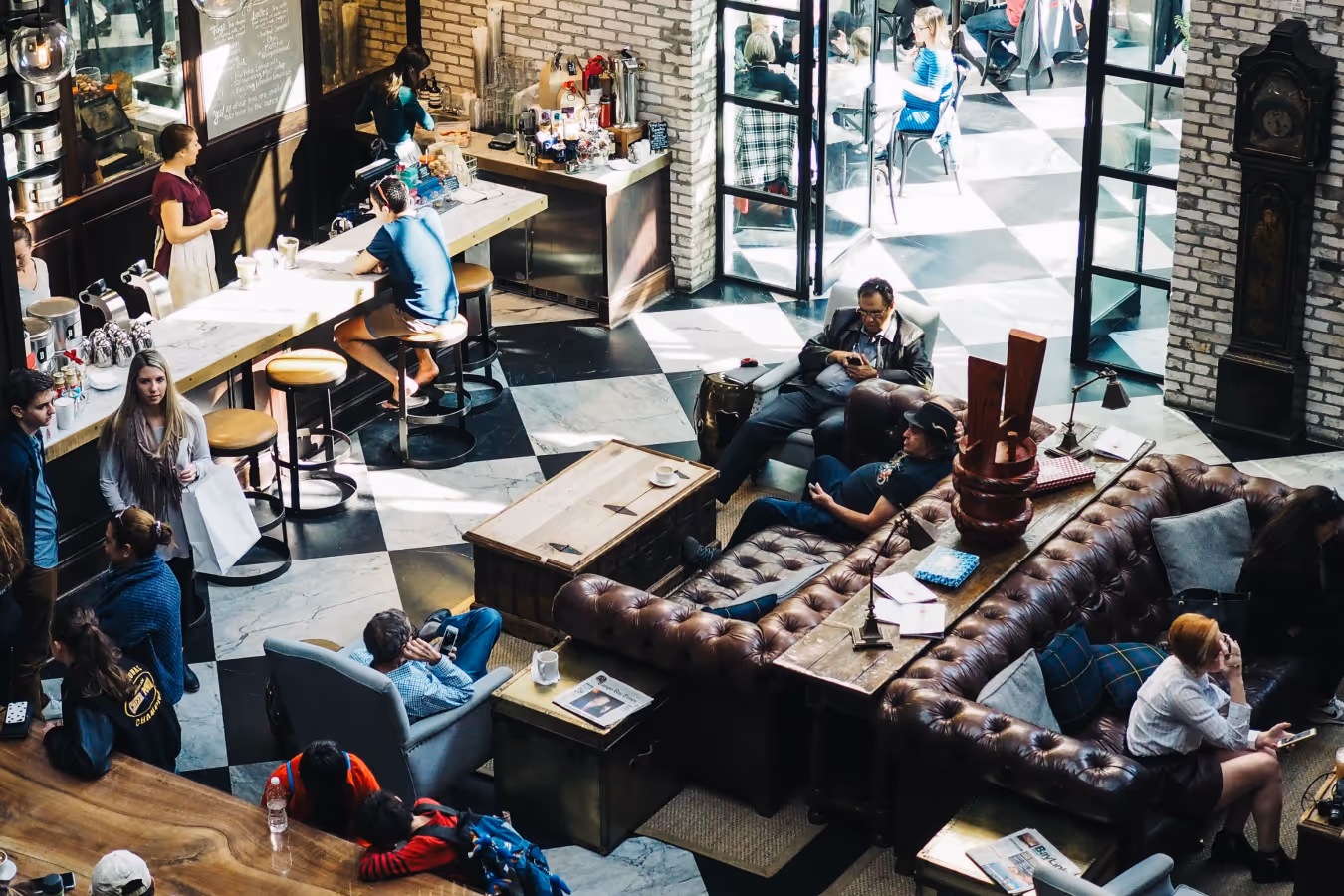 People sitting and standing in a busy cafe with checkered floor, leather sofas, and wooden tables.