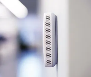 Close-up of a modern white wall-mounted IoT device with perforated grille in a blurred interior setting.