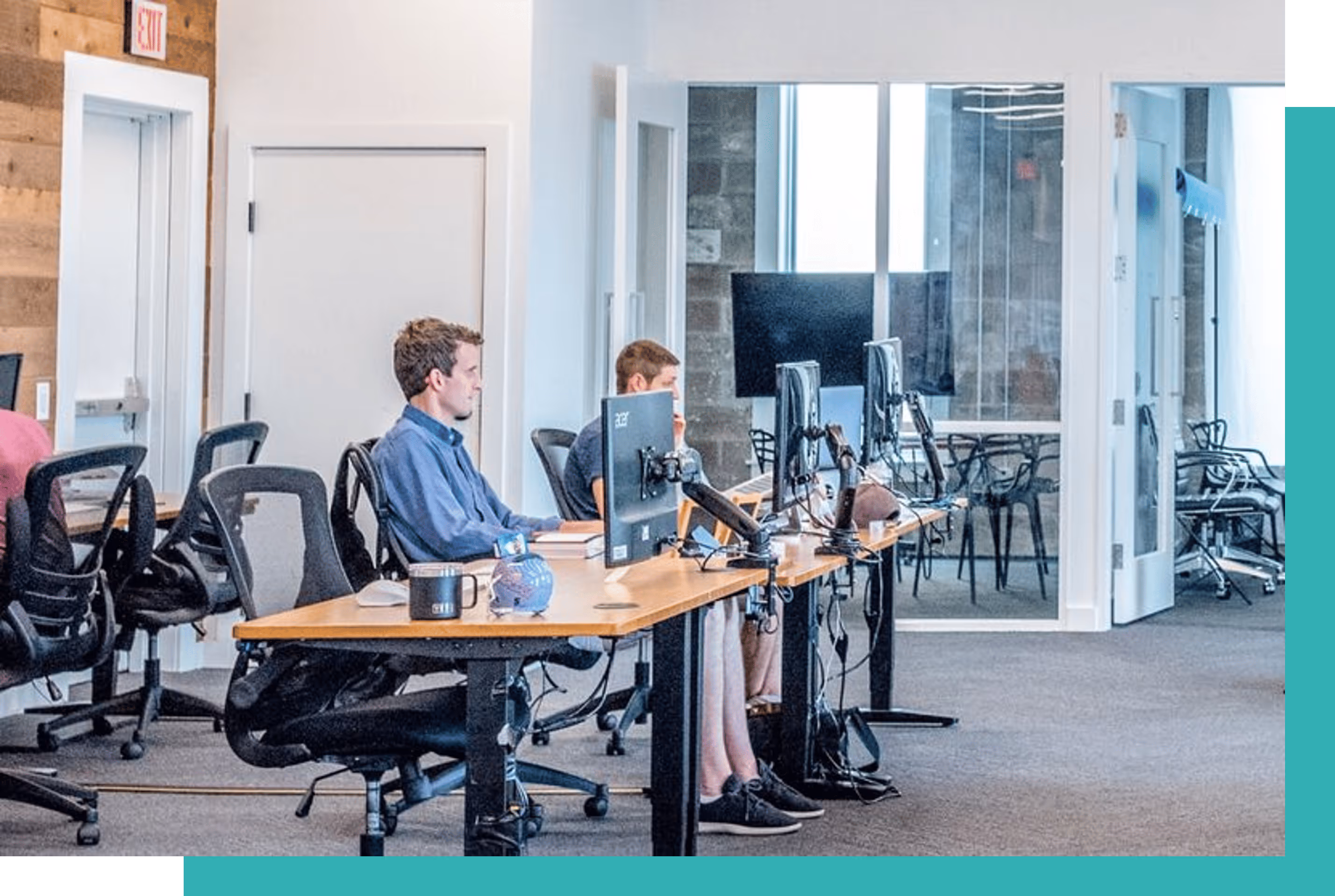 Two men working on desktop computers at a long shared office desk in a modern, open-plan workspace.