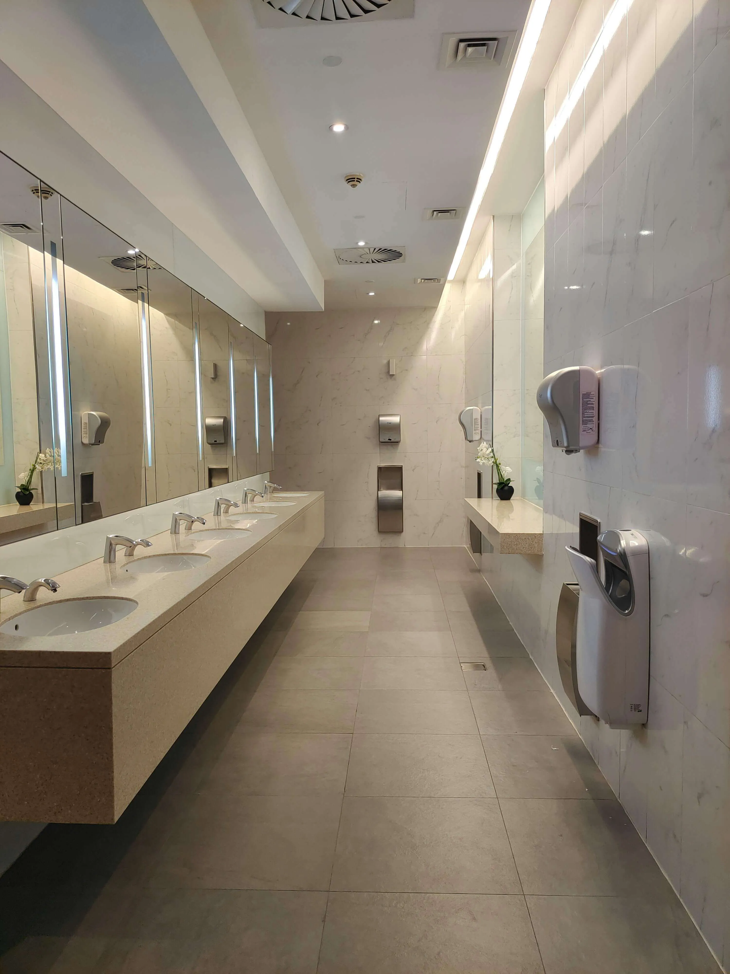 Modern public restroom with multiple sinks, mirrors, hand dryers, soap dispensers, and a marble wall.