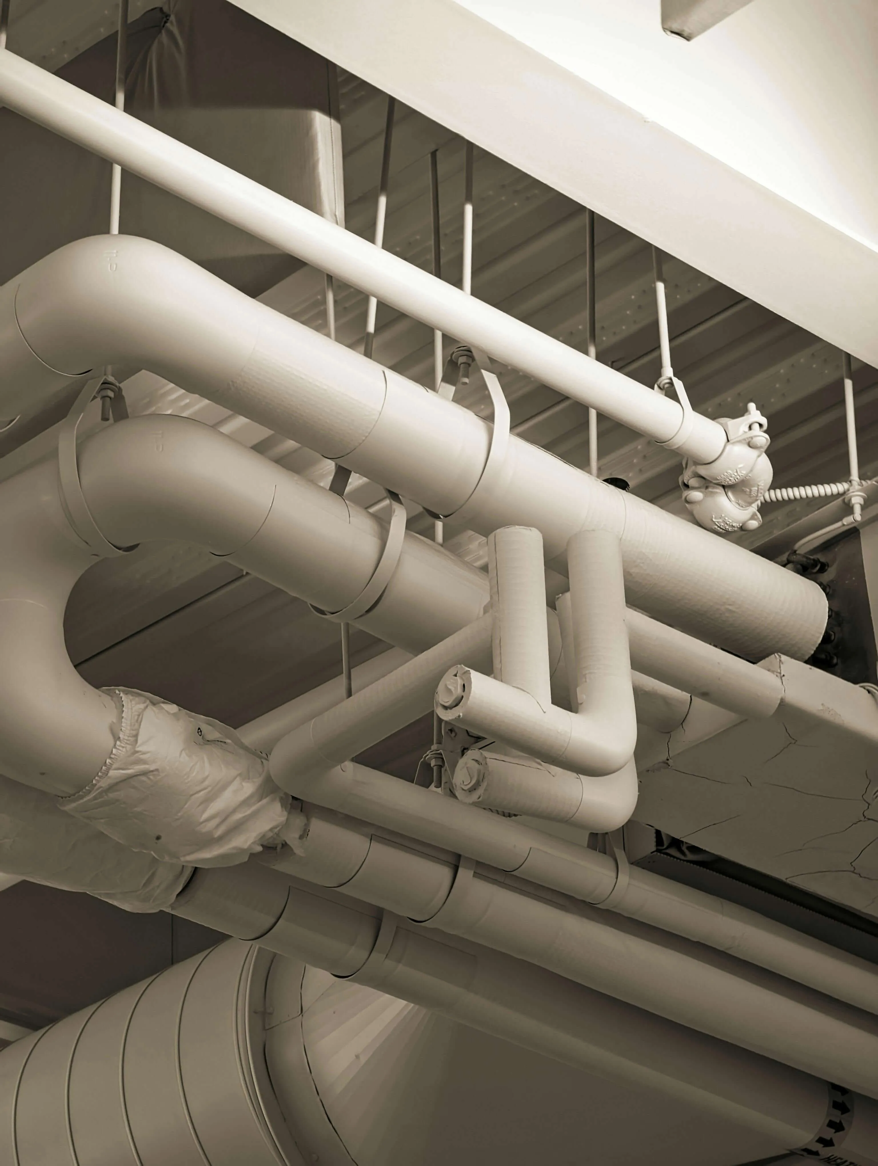 Ceiling with multiple interconnected industrial pipes and ductwork in beige tones.