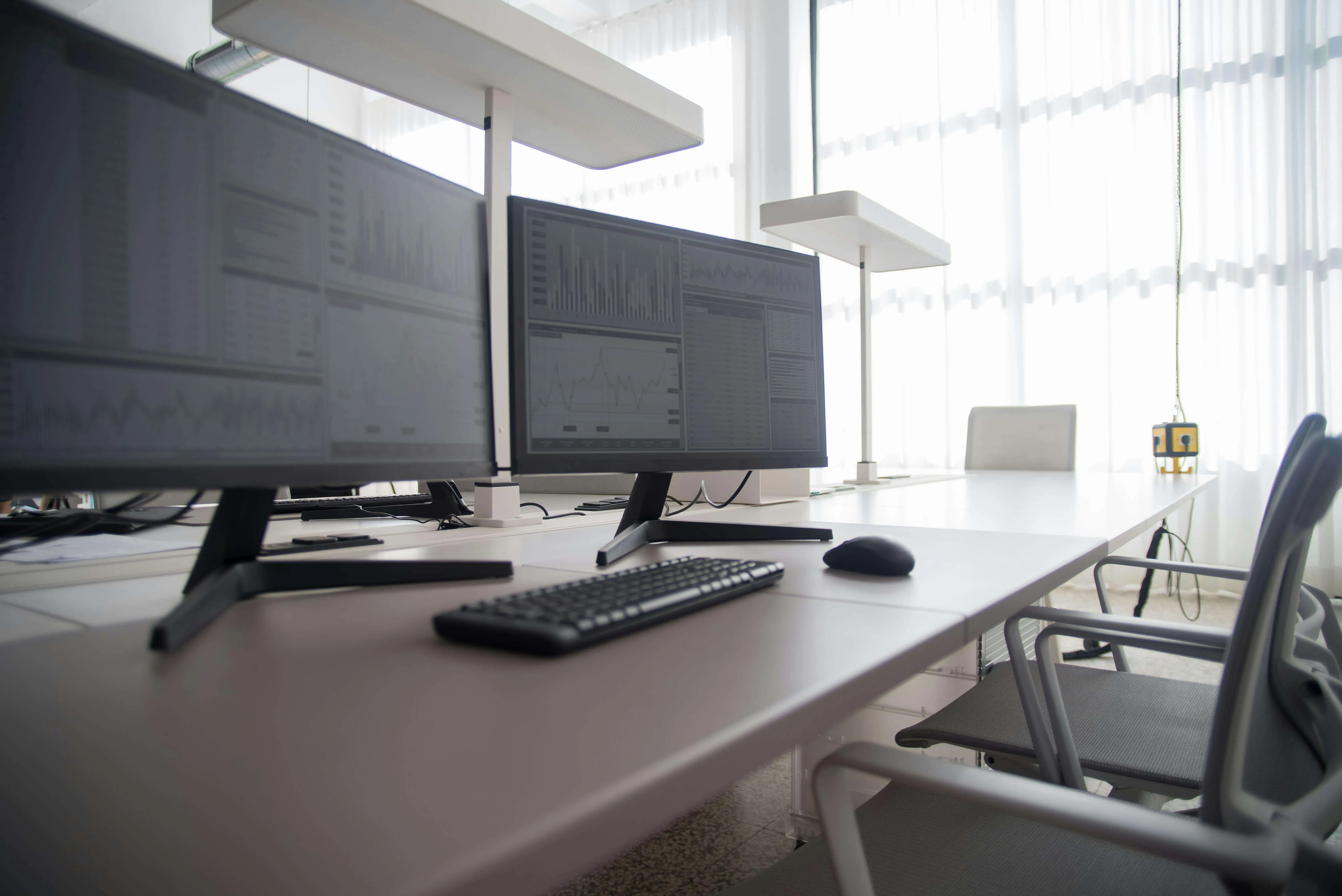 Office desk with dual monitors displaying graphs, keyboard, mouse, and office chairs near large windows.