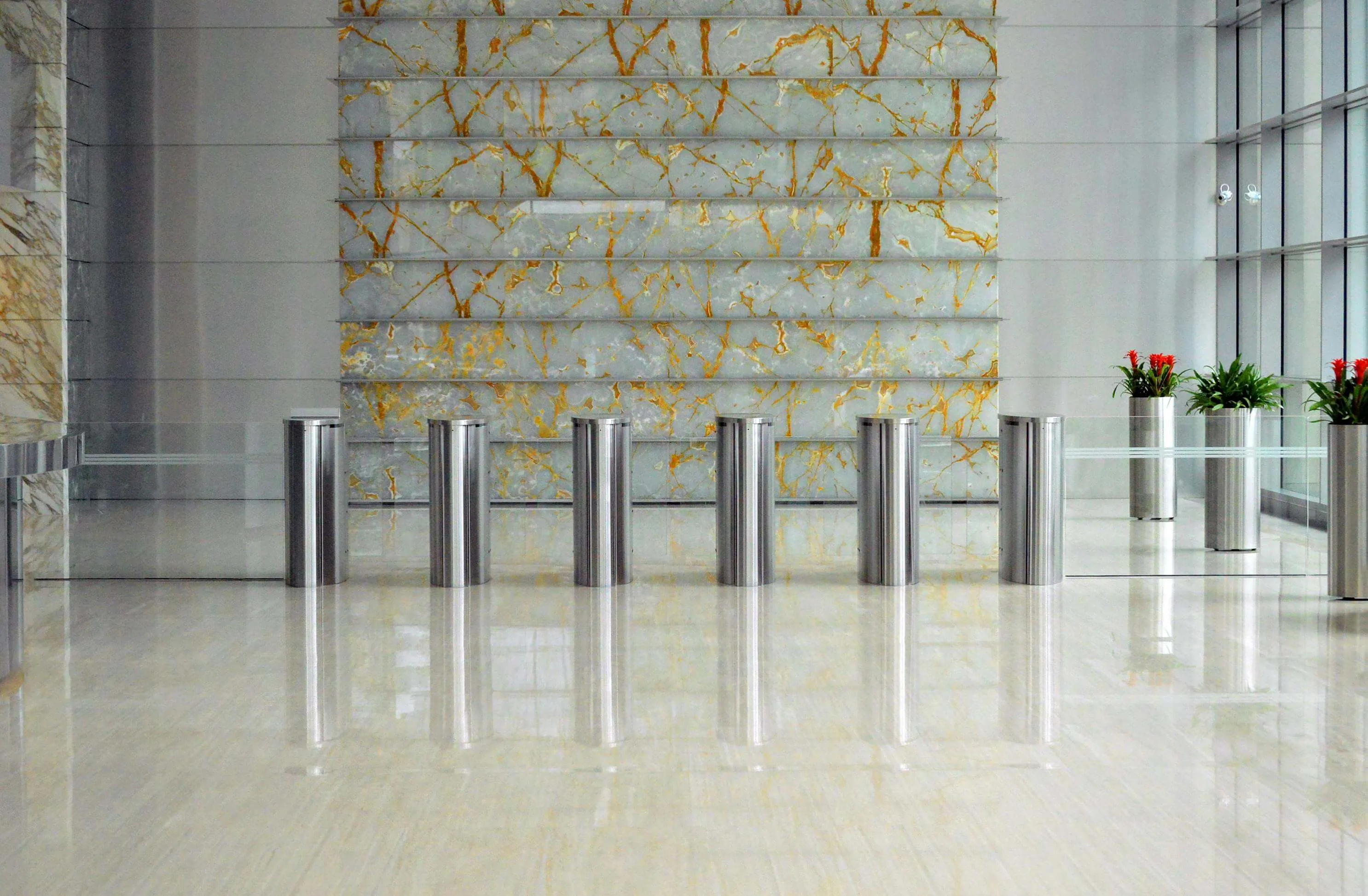 Modern building lobby with a marble wall, stainless steel turnstiles, and potted plants with red flowers.