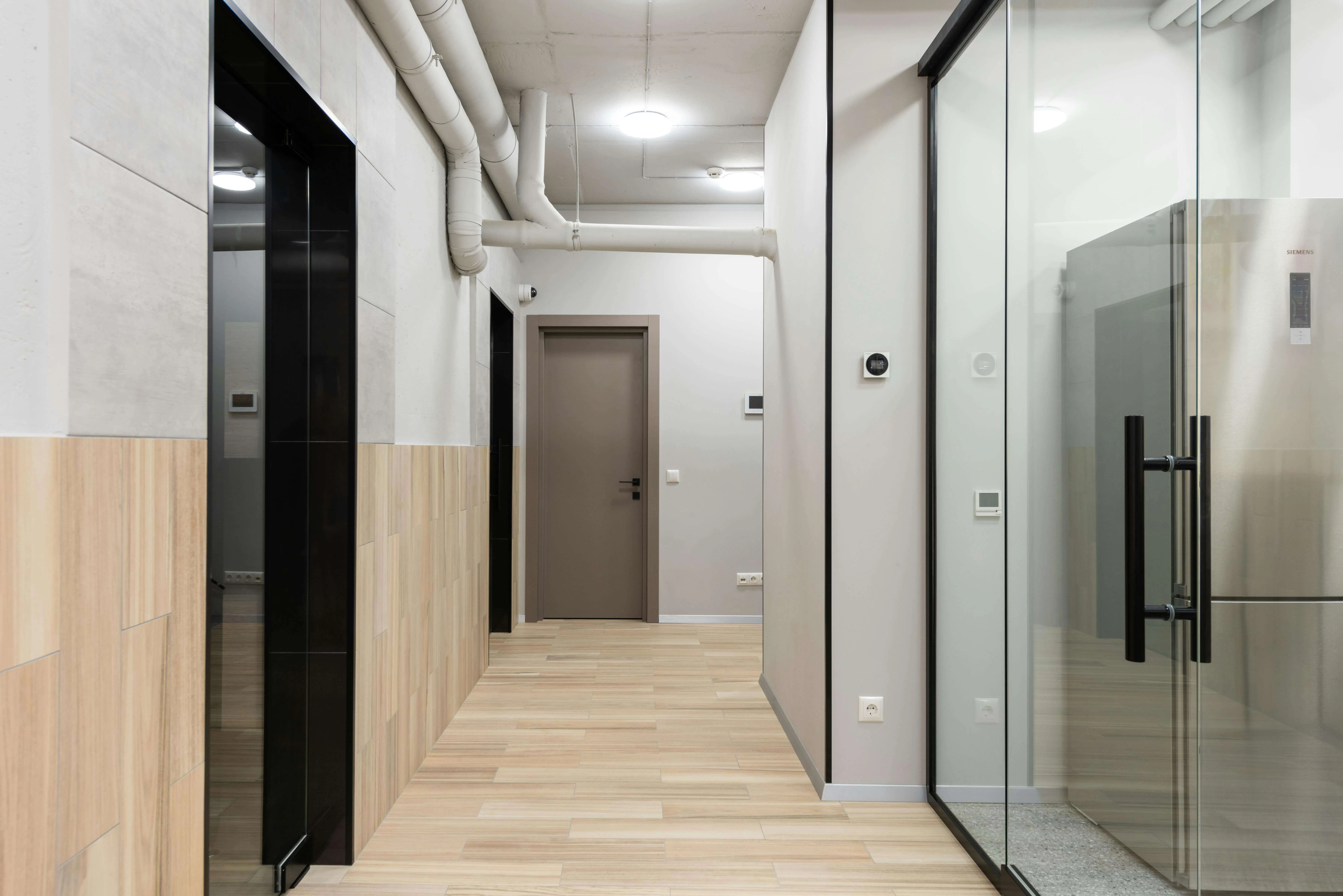 Modern hallway with light wood floor, closed door, exposed pipes, and glass wall showing a refrigerator.