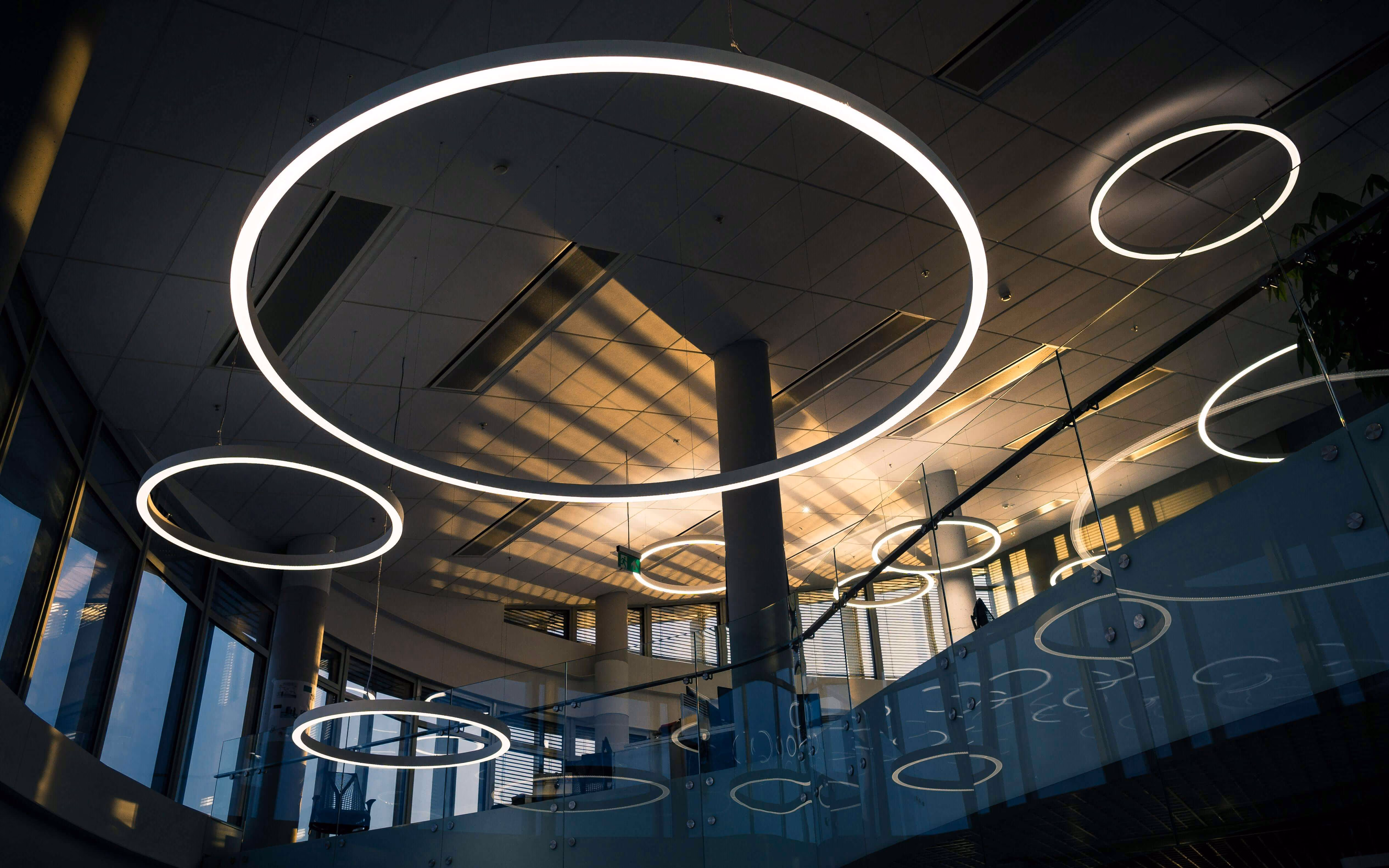 Modern office ceiling with large circular hanging LED lights and reflections on glass panels.