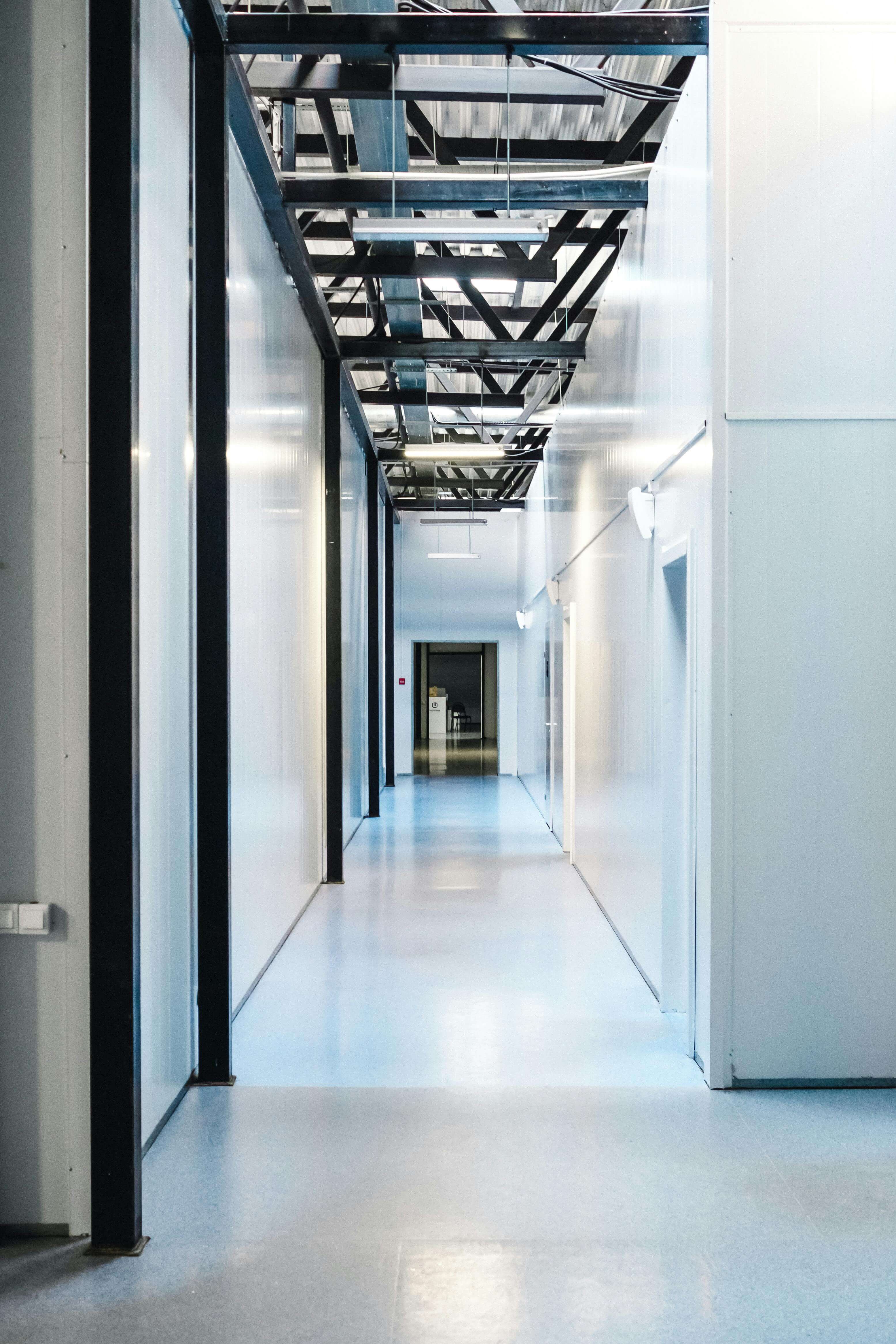 Empty industrial hallway with white walls, blue floor, exposed ceiling beams, and doors on the right.