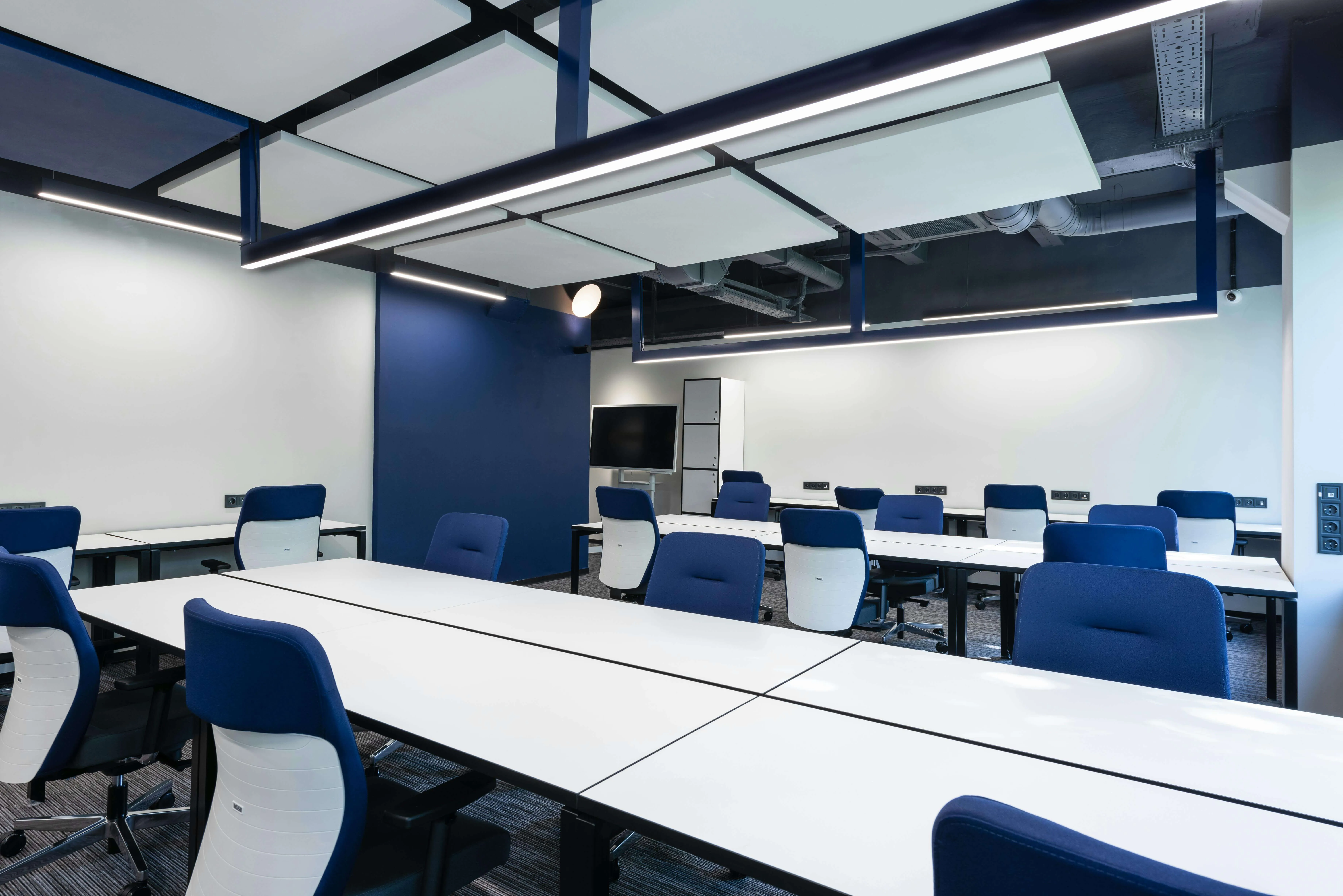 Modern empty office space with white desks and blue office chairs under ceiling panels with lights.
