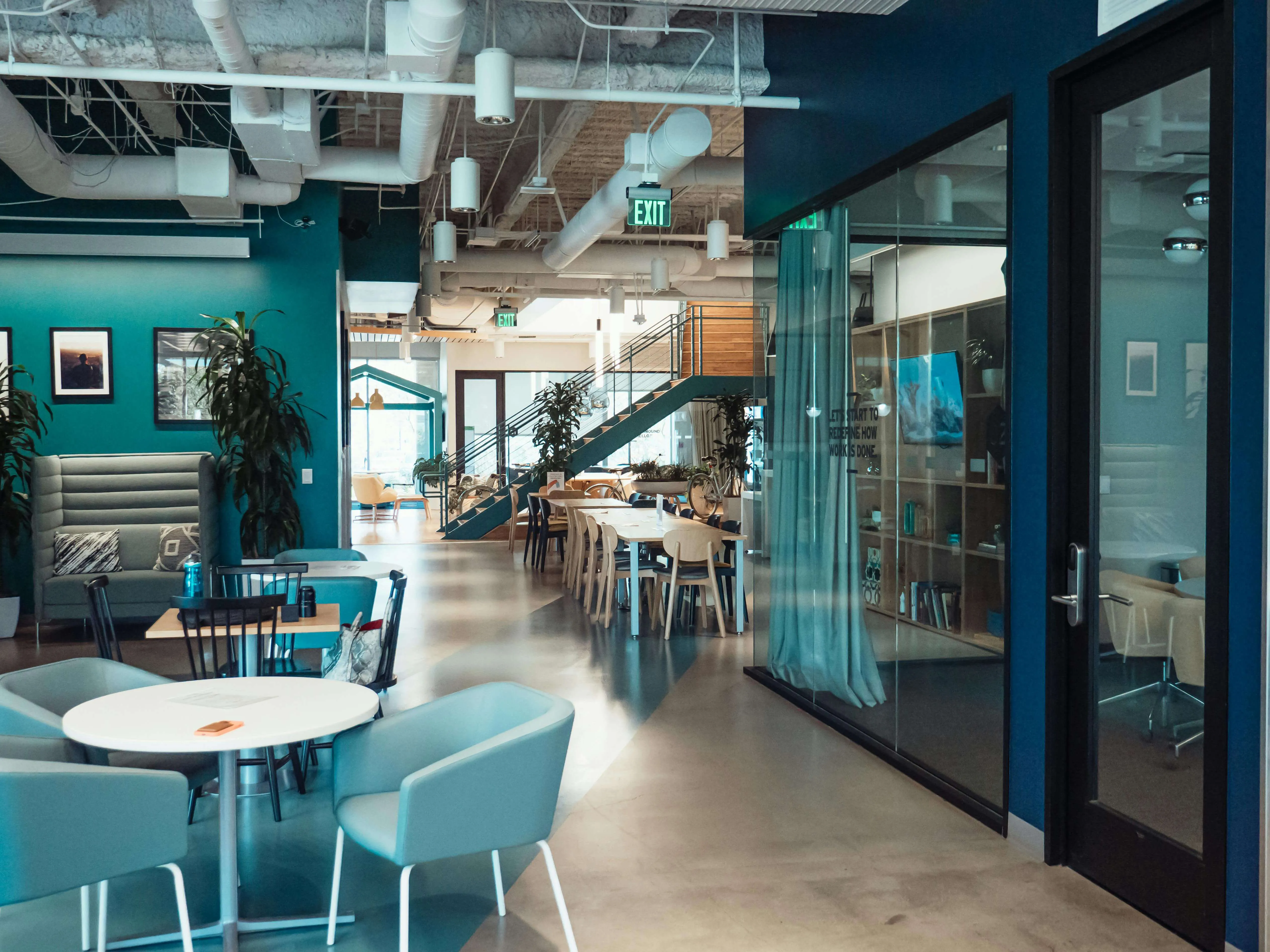 Modern office lounge with blue chairs, tables, plants, staircase, and glass-walled meeting room.