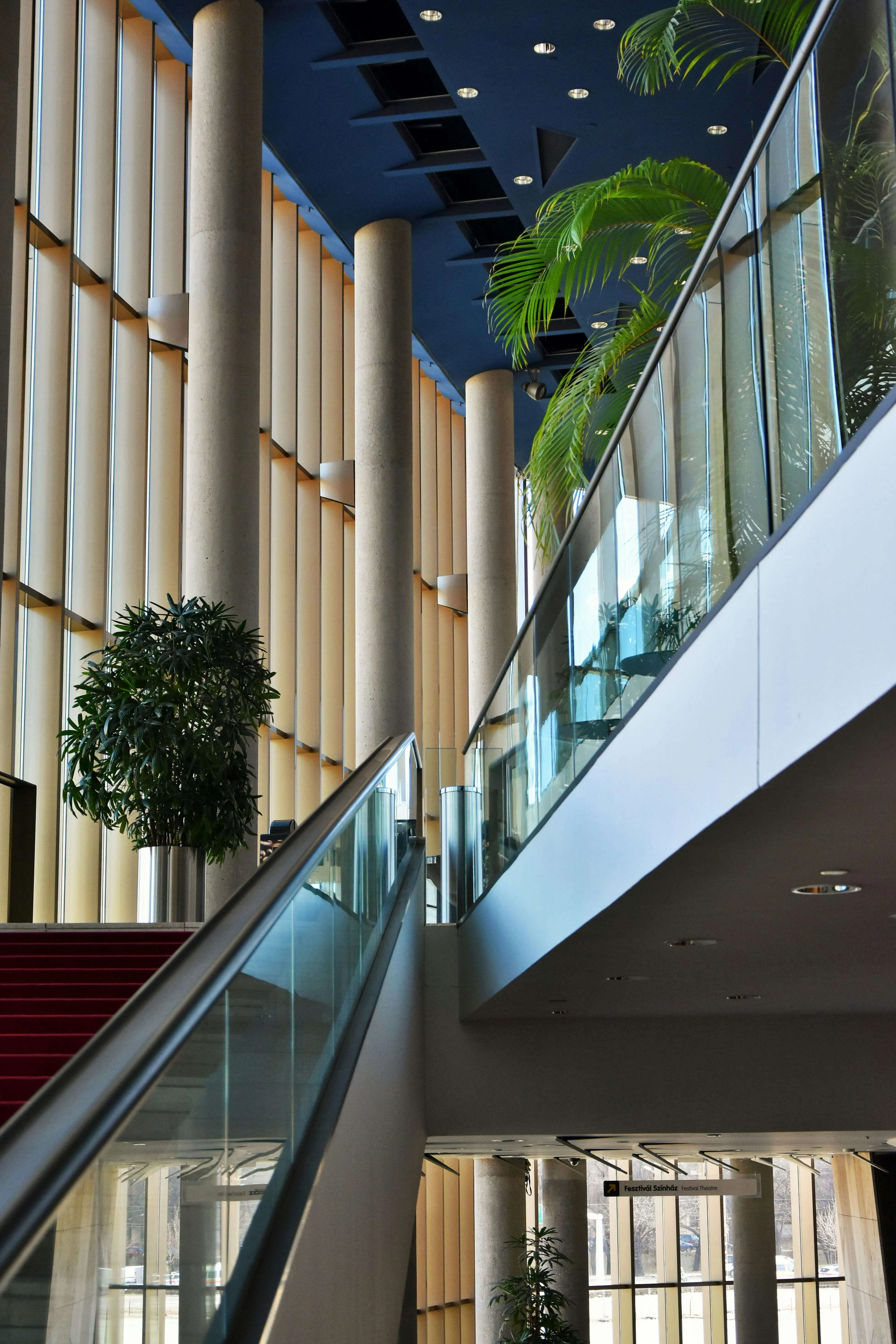 Interior showing a stairway with glass railing, tall columns, large windows, and potted plants.