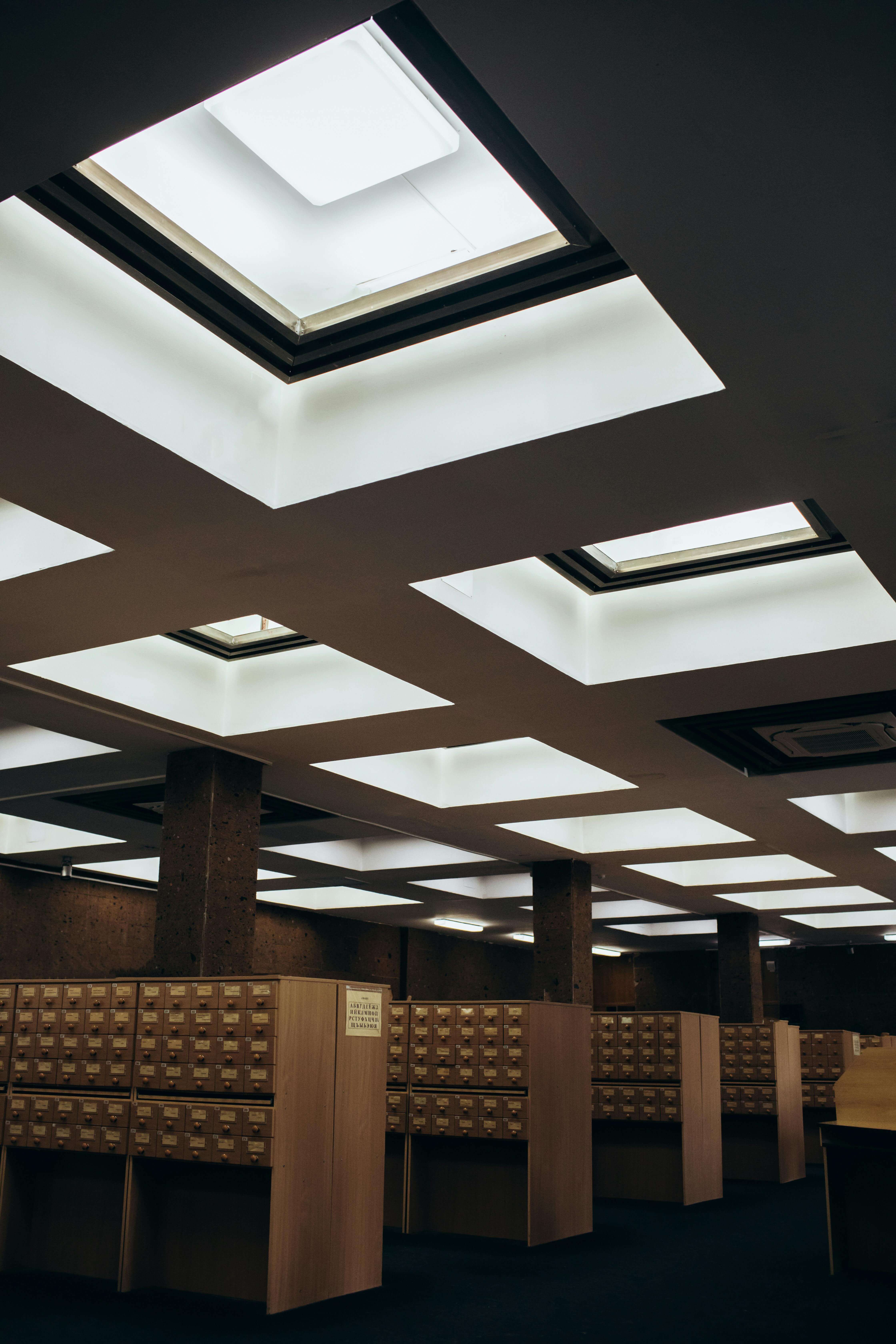 Rows of wooden card catalog cabinets under ceiling skylights in a dimly lit library room.