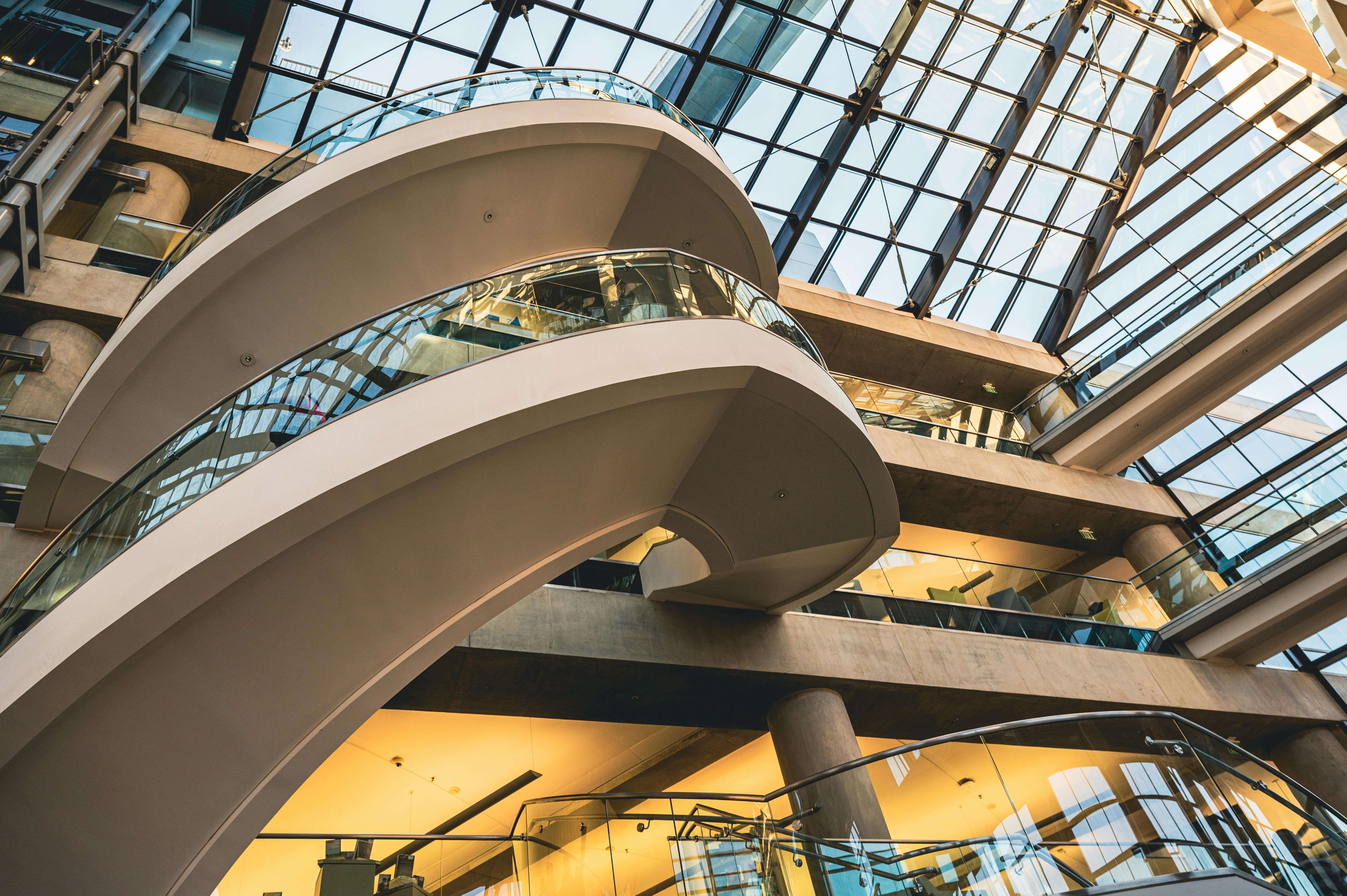 Curved modern staircase inside a building with glass ceiling and concrete pillars.