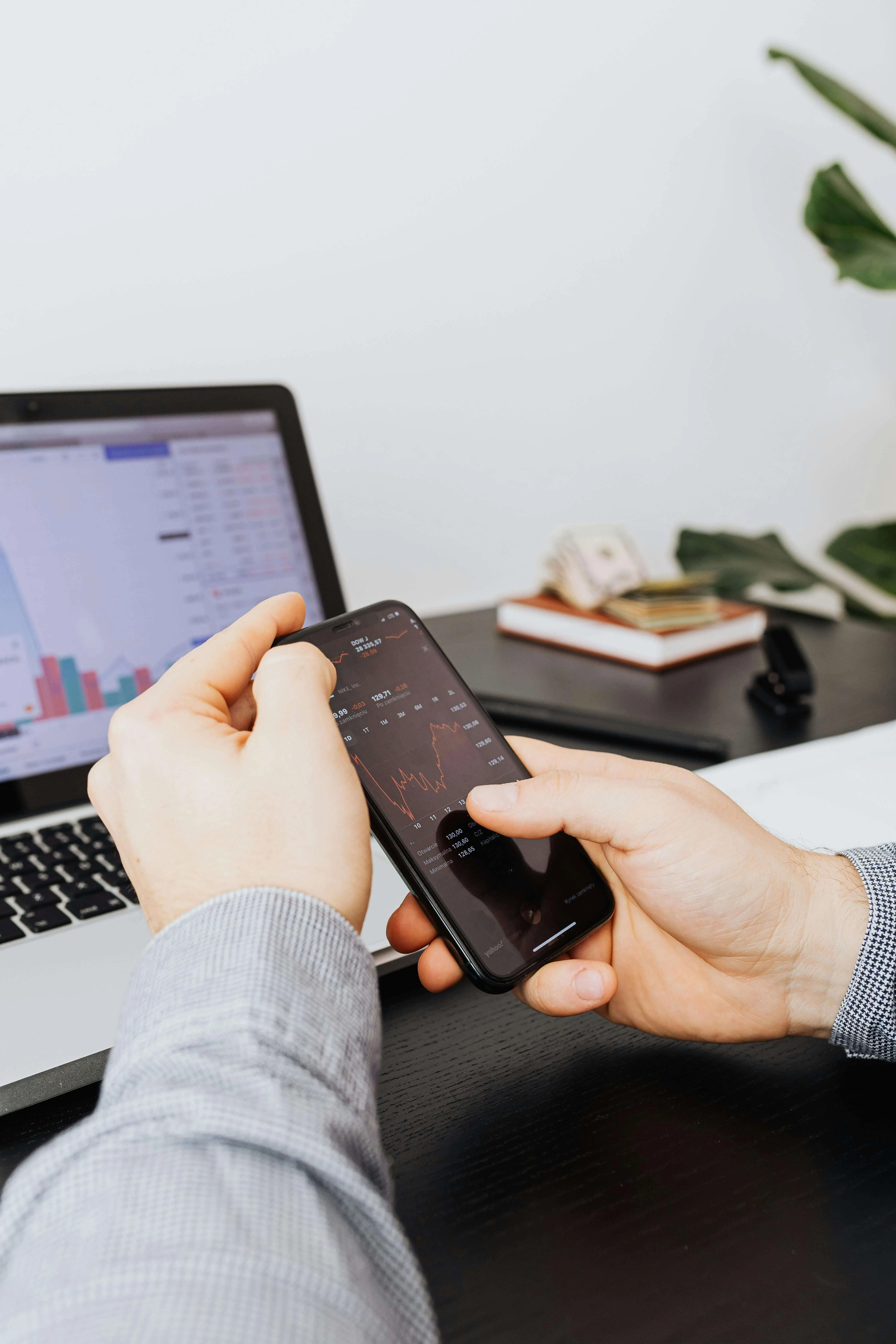 Person checking stock market charts on a smartphone with a laptop displaying graphs on a desk.