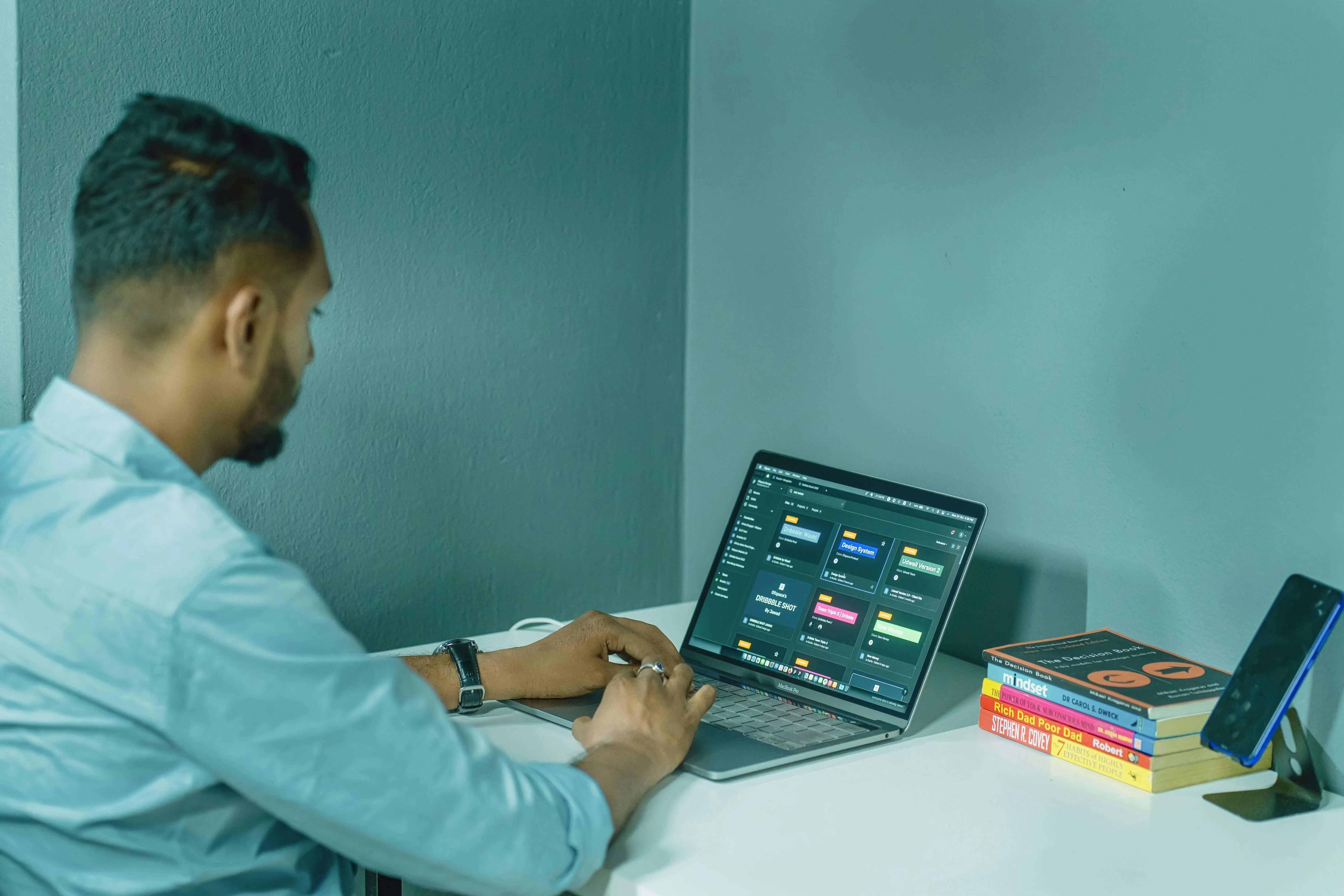Man working on a laptop at a white desk with a stack of books and a phone on a stand.