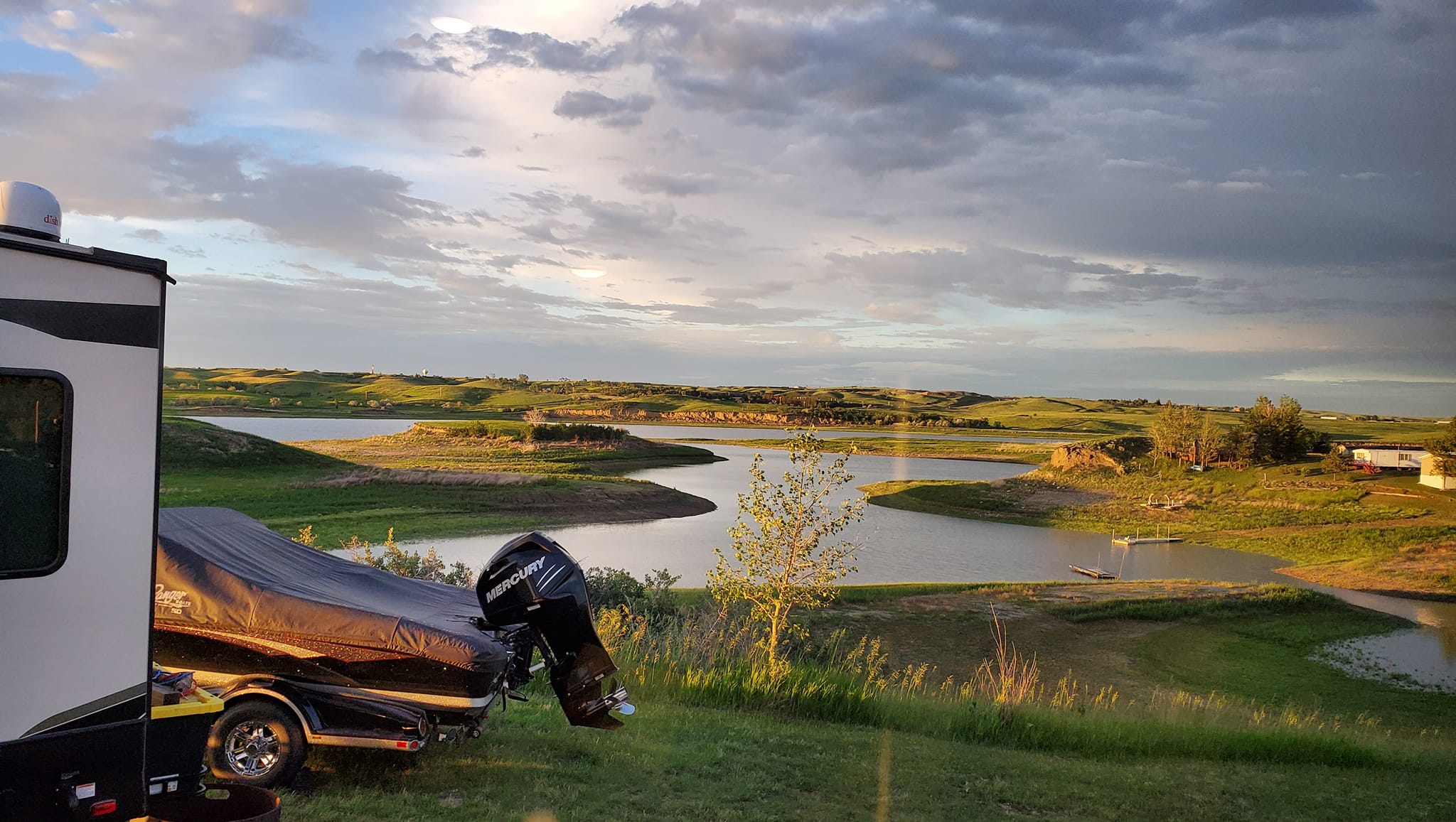 Covered motorboat on trailer next to recreational vehicle overlooking a winding river and green rolling hills under a partly cloudy sky during sunset.