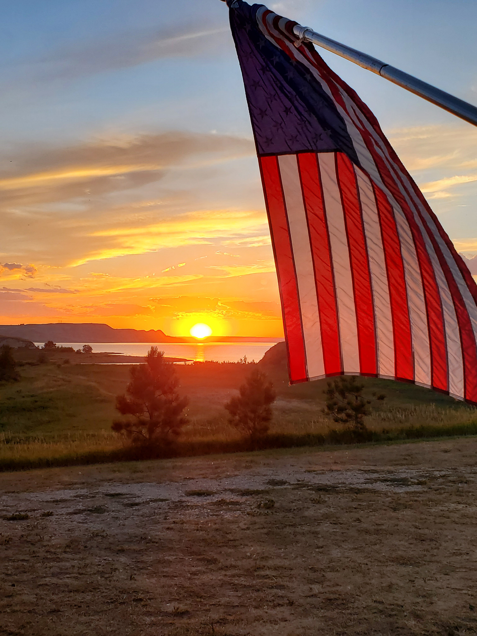 American flag waving in the foreground with a sunset over a lake and hills in the background.