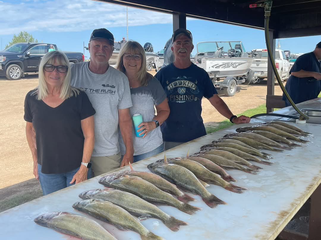 Four adults standing behind a table displaying a row of freshly caught fish outdoors near boats and trucks.