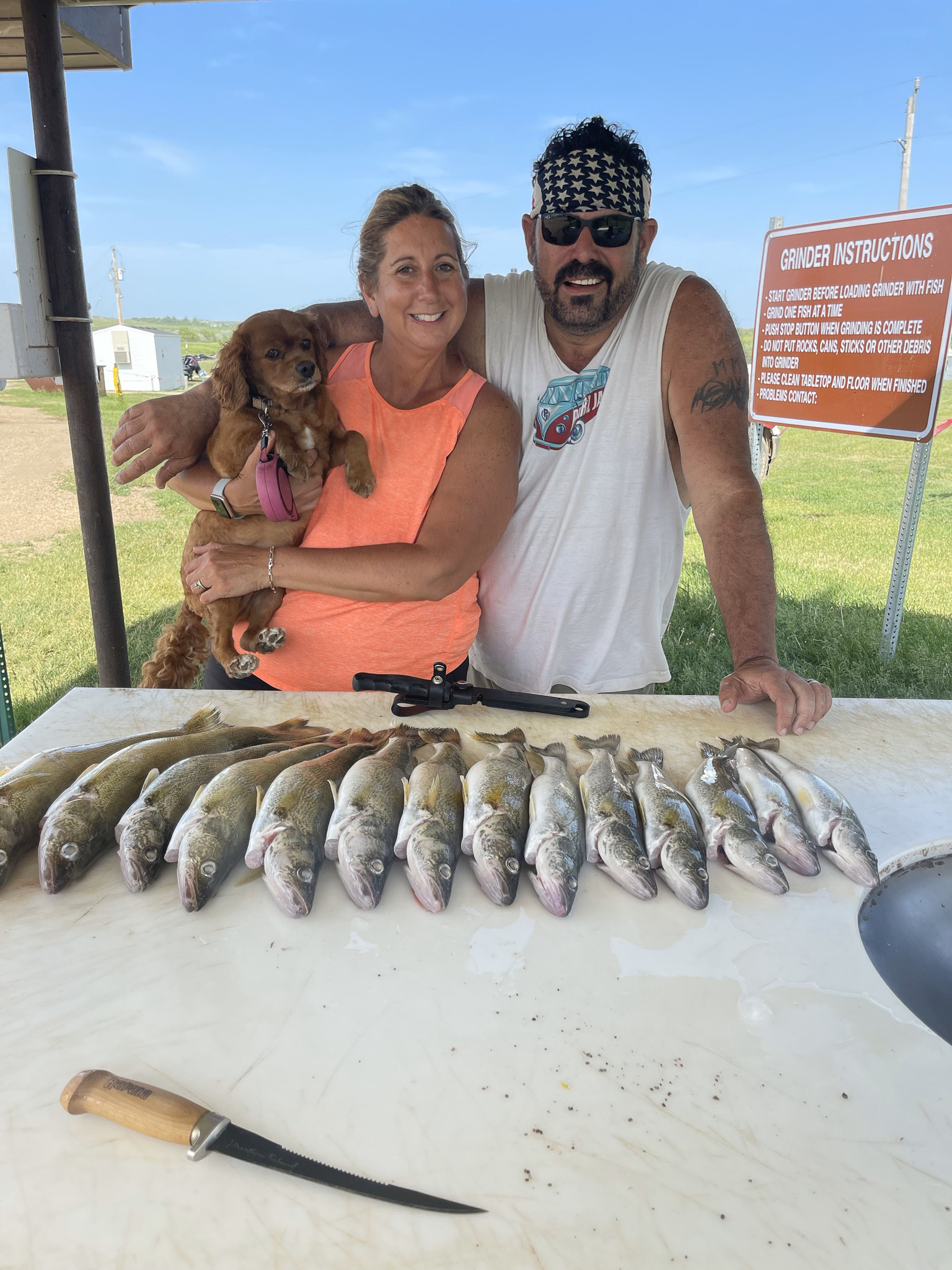 Smiling man and woman standing behind a table with twelve fish laid out and a fillet knife, woman holding a small brown dog.