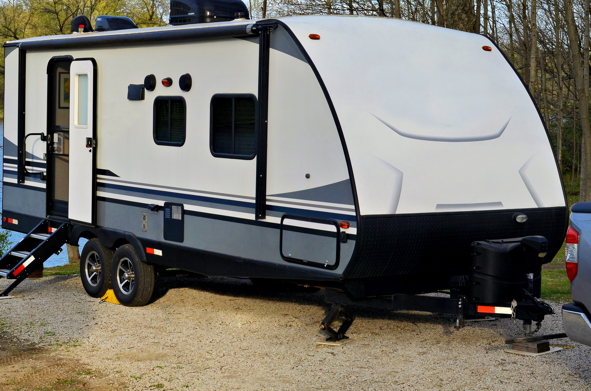 White and gray travel trailer parked on gravel near trees with open door and extended steps.