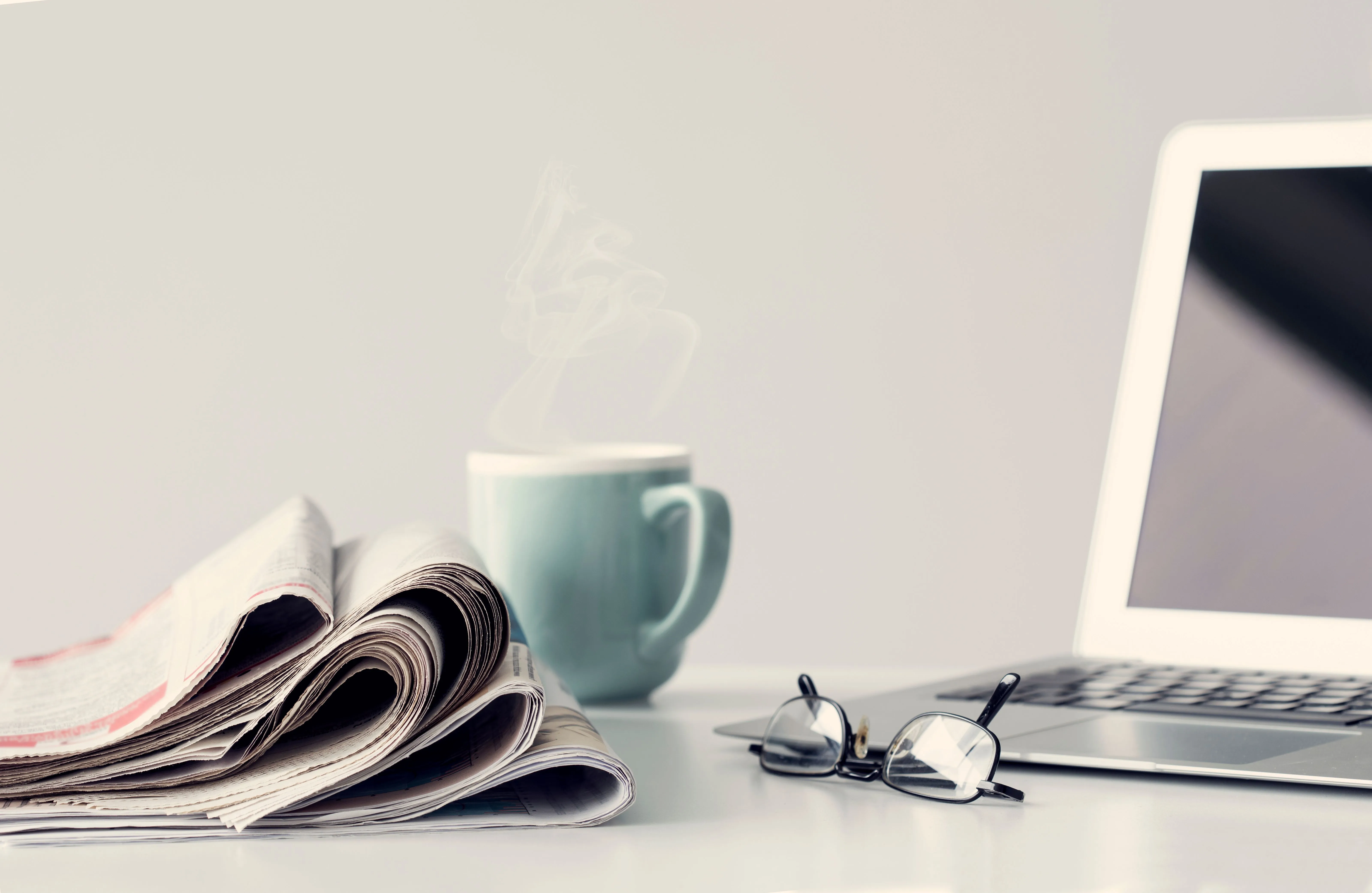 Newspaper, coffee mug, glasses, and laptop on a minimalist workspace