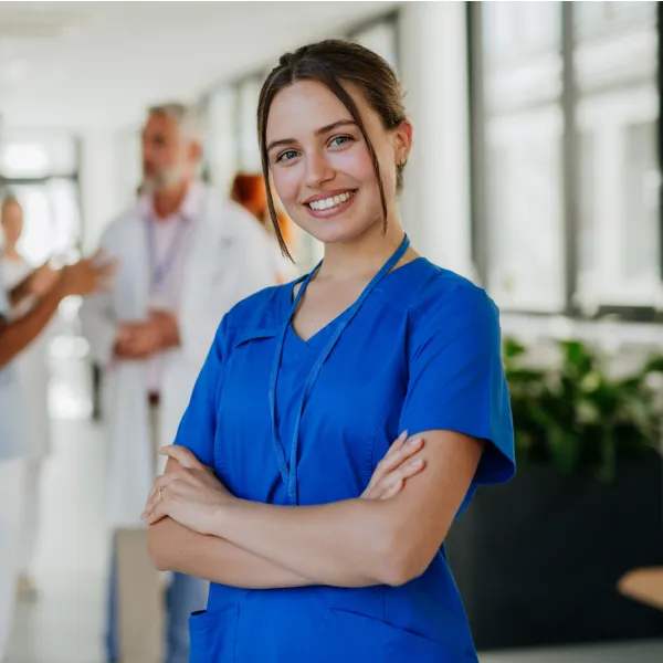 Smiling young female nurse wearing blue scrubs with arms crossed in a hospital corridor with blurred medical staff in the background.