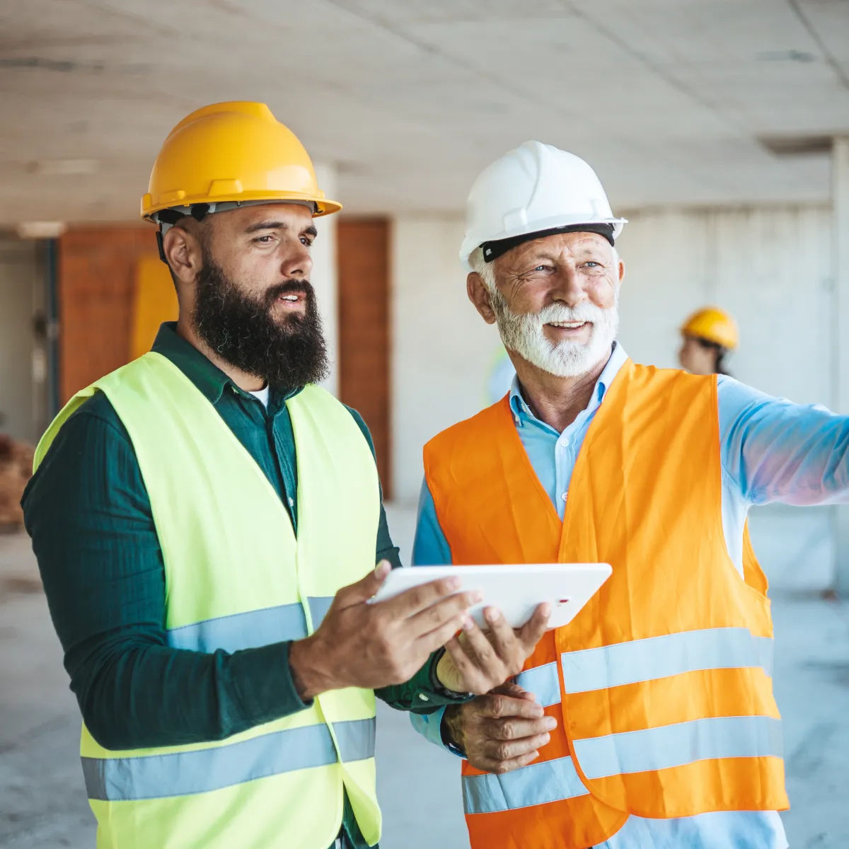Two construction workers wearing helmets and reflective vests discussing plans using a tablet on a construction site.