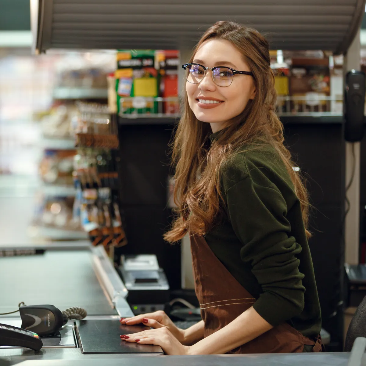 Smiling young female cashier with glasses and long hair standing at checkout counter in a store.