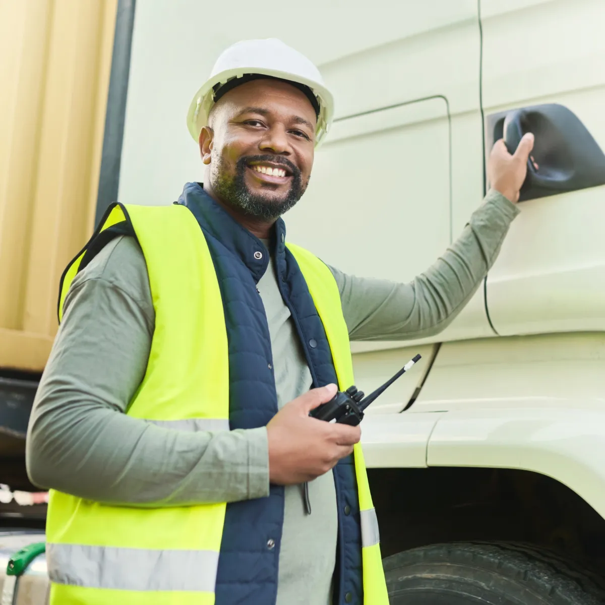 Smiling man wearing a white hard hat and reflective vest holding a walkie-talkie and opening the door of a large truck.