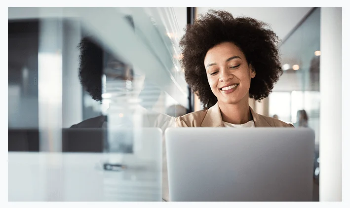 Smiling woman with curly hair working on a laptop in a modern office setting.