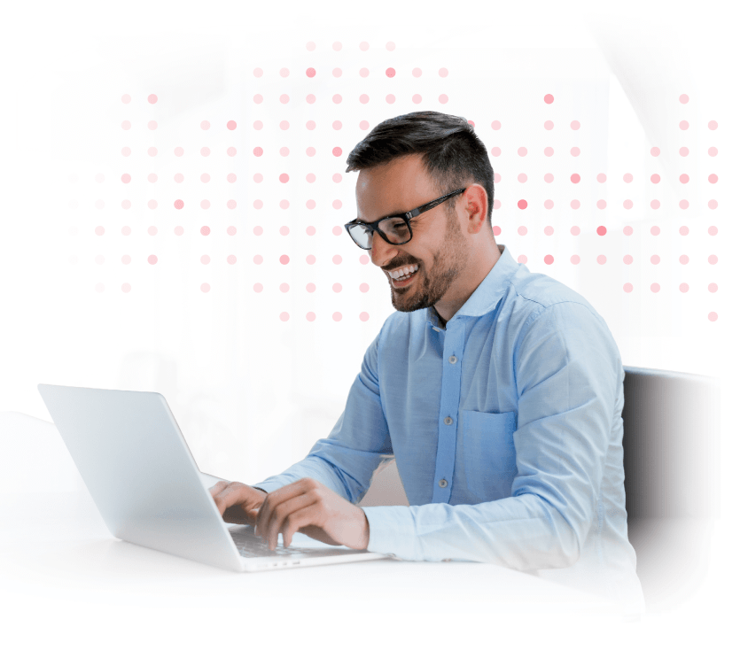 Man with glasses smiling while typing on a laptop at a white desk.