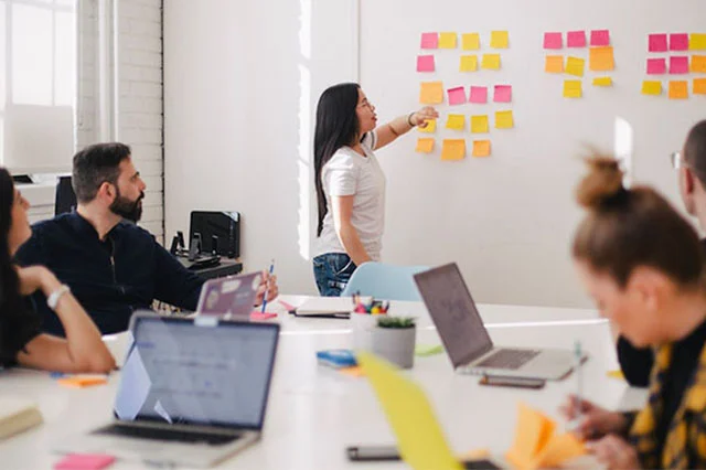 Woman points to colorful sticky notes on wall while colleagues sit at table with laptops and papers.