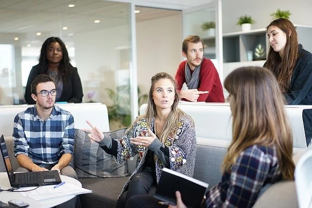 Five diverse young adults in casual clothes having a discussion in a modern office lounge.