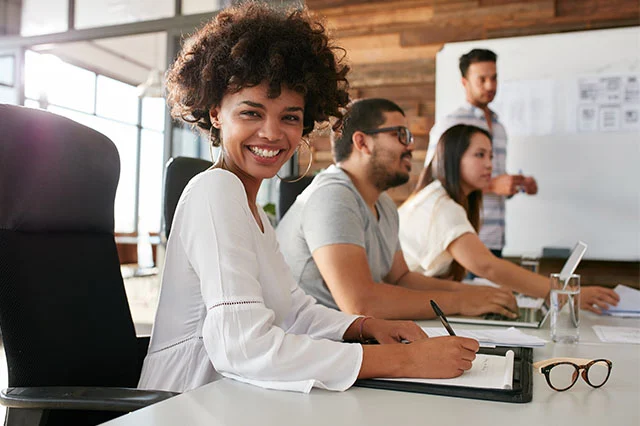 Smiling woman with curly hair writing while colleagues work and discuss in an office meeting.