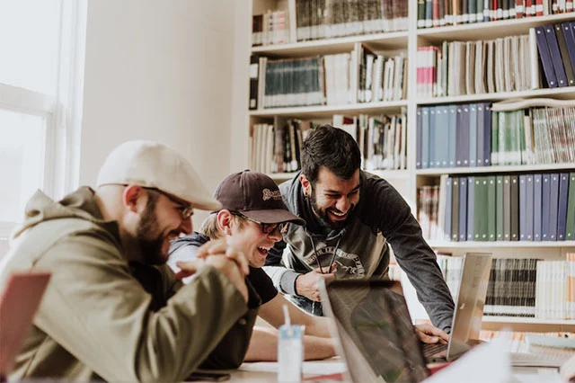 Three men laughing and working together at laptops in a library with bookshelves in the background.
