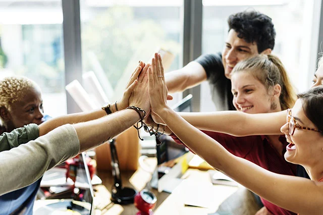 Group of diverse coworkers sitting at a table giving a high five in a bright office.