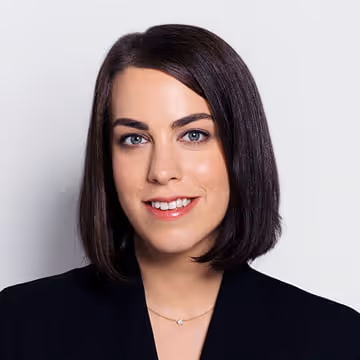 Portrait of a woman with straight dark hair, blue eyes, wearing a black blazer and a delicate necklace.