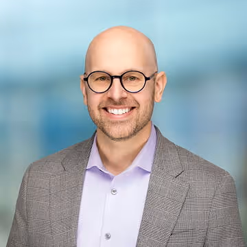Smiling bald man with glasses wearing a gray blazer and light purple shirt on a blue blurred background.