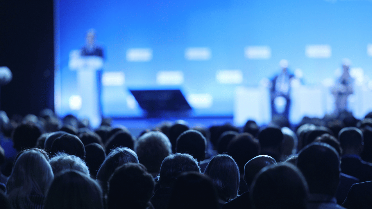 Audience watching a speaker on stage at a conference or event with a blue backdrop.