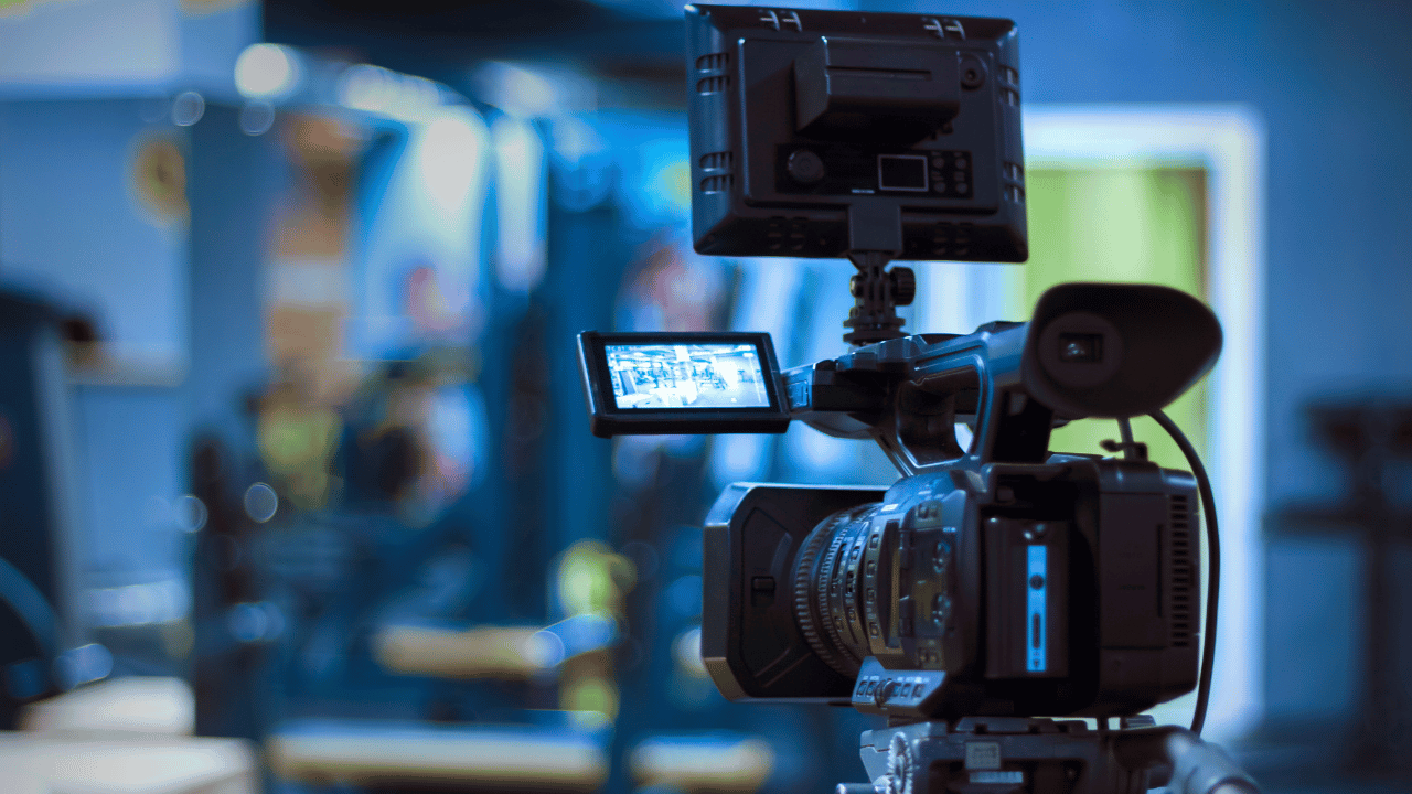 Professional video camera on a tripod with its LCD monitor flipped out in a blurred indoor setting.