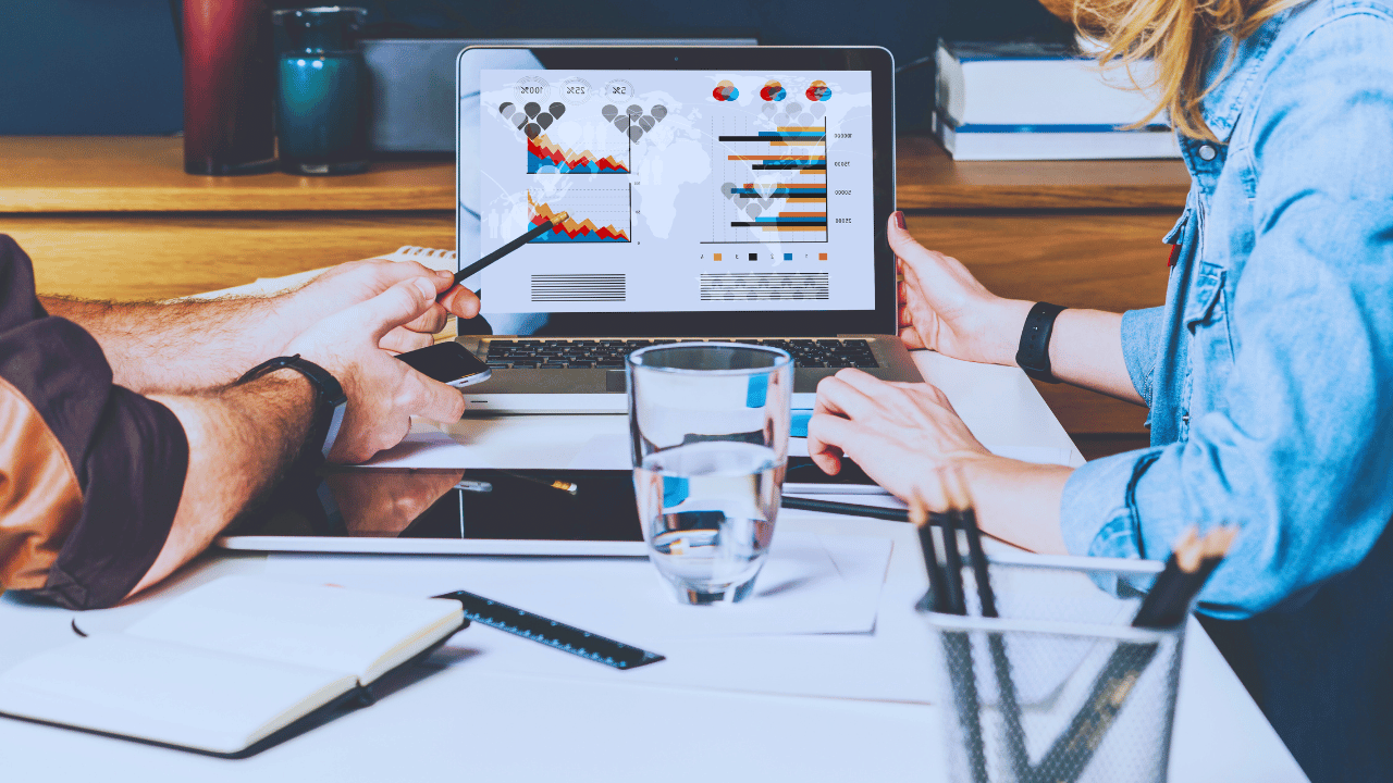 Two people discussing data charts and graphs displayed on a laptop screen at a desk with office supplies.
