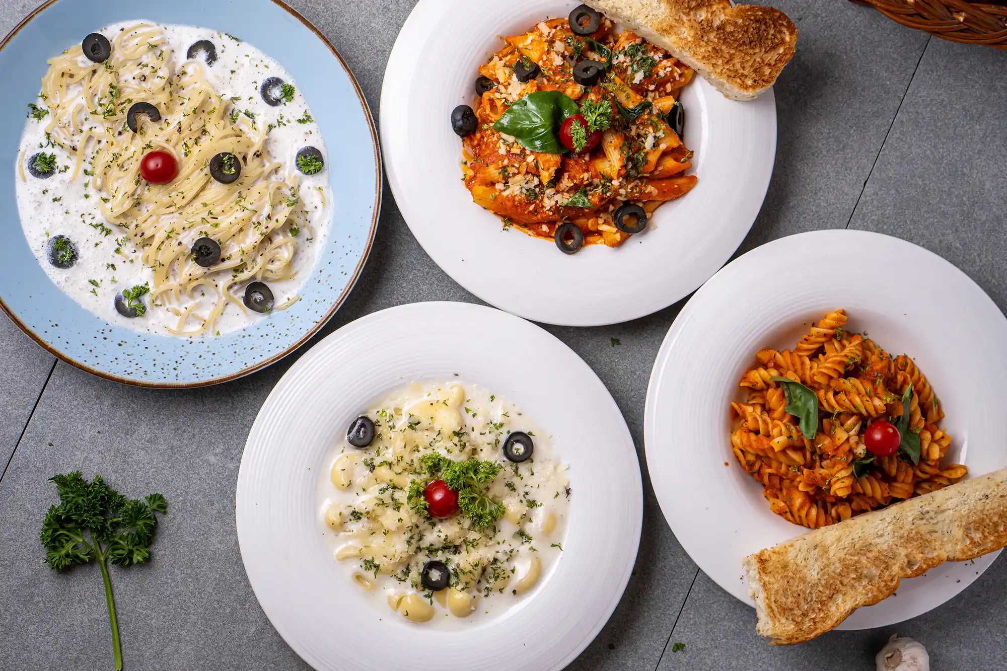 Four plates of different pastas: creamy linguine with olives and cherry tomato, penne with tomato sauce, fusilli with tomato sauce and basil, and macaroni in creamy sauce with olives and cherry tomato, served with bread.
