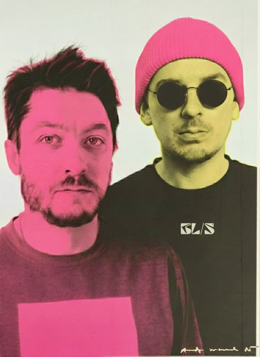 Two men posing side by side, one with a beard and tousled hair, the other wearing round sunglasses and a pink beanie.