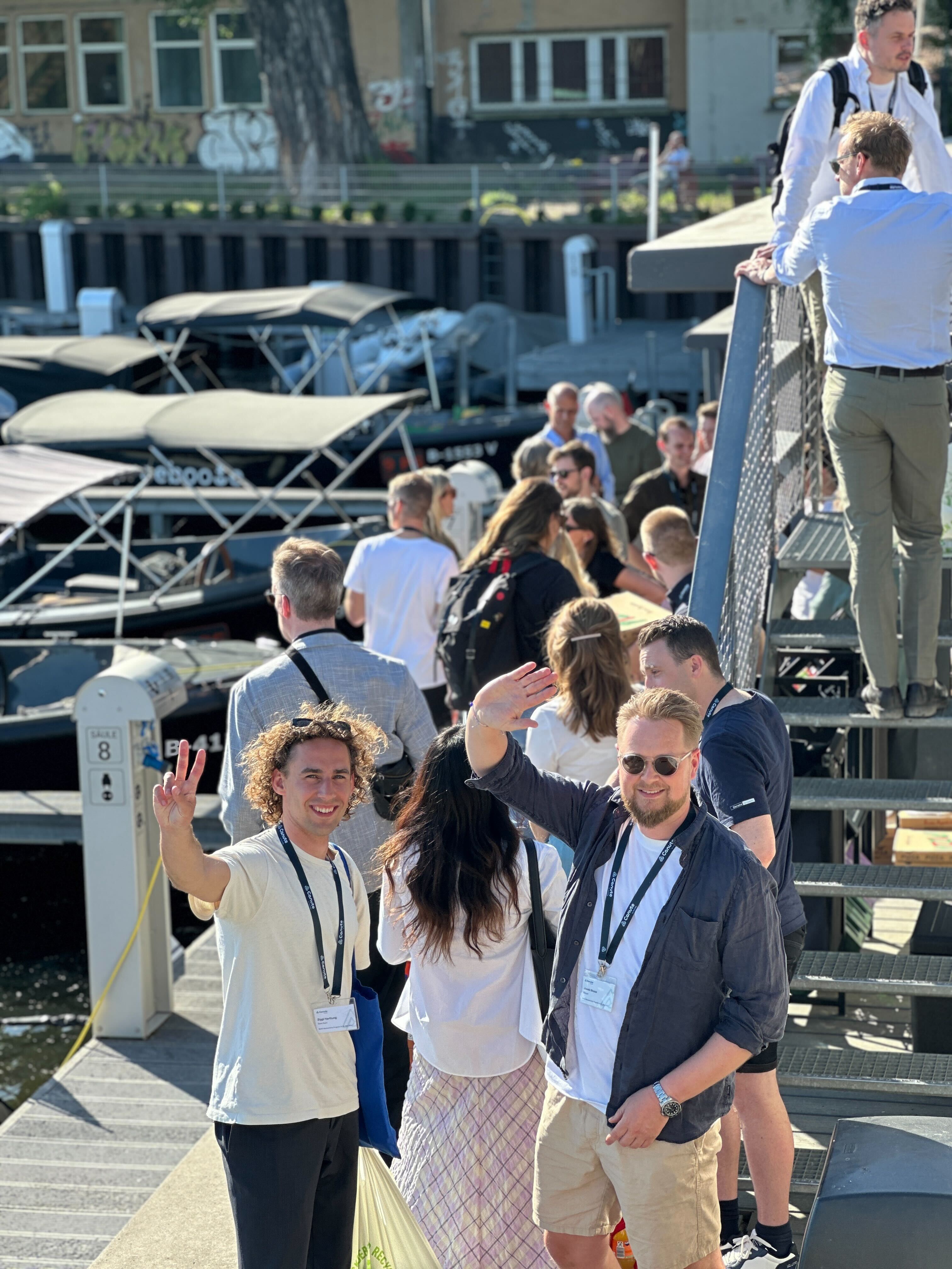 Group of people on dock near boats with two men waving and smiling at the camera in sunlight.