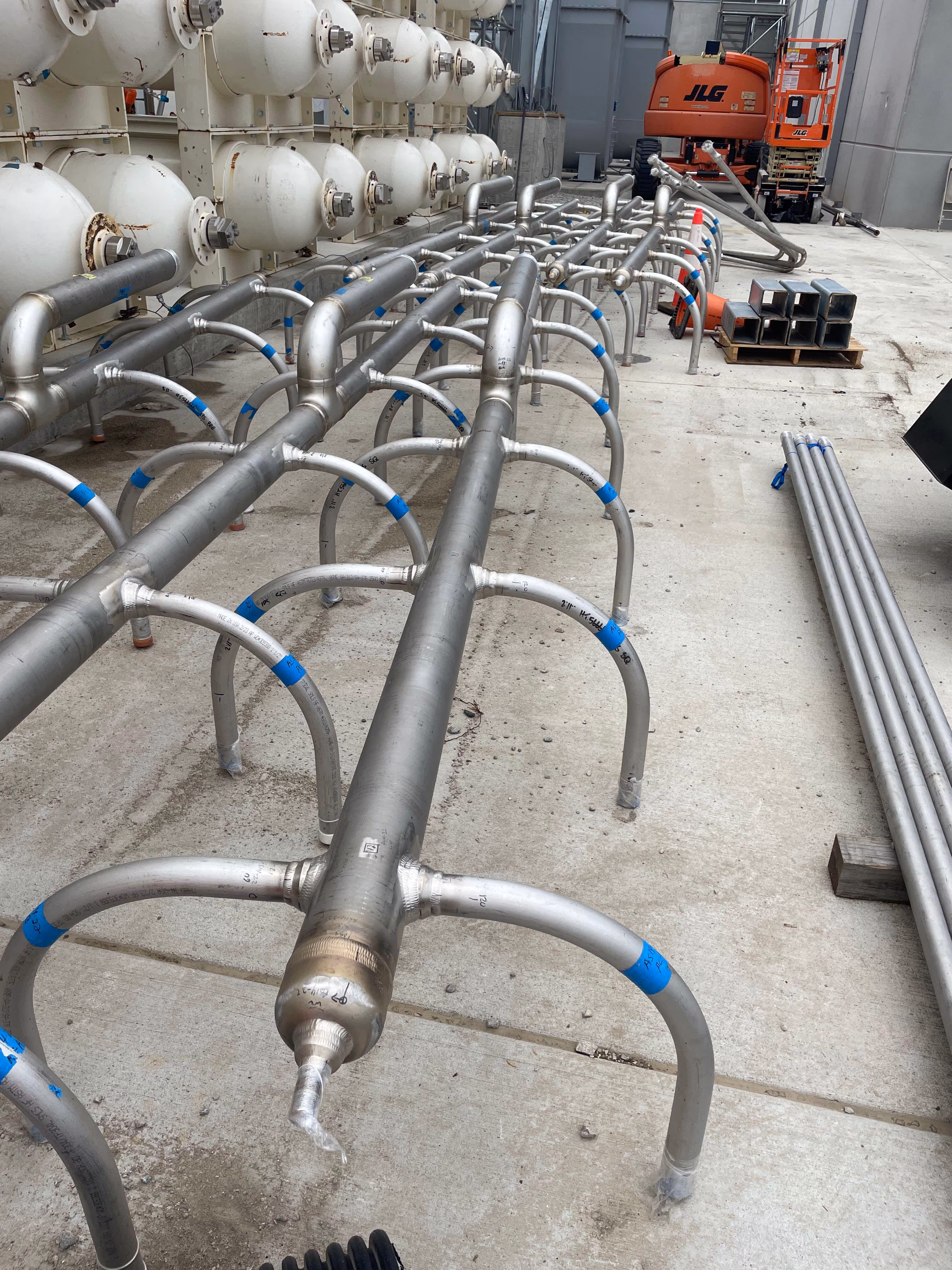 Industrial stainless steel piping system with curved support legs on a concrete floor inside a facility.