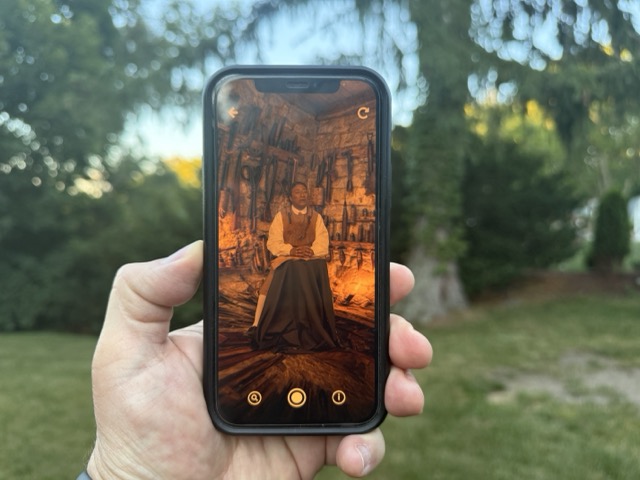 Hand holding a smartphone outdoors, screen is displaying a historical scene of a man in old-fashioned clothing seated in a rustic stone room with tools hanging on the walls.
