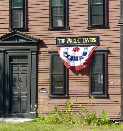 Exterior of The Wright Tavern, a brown wooden building with three upper windows and two lower windows; a patriotic red, white, and blue bunting hangs below the tavern sign, and a black door with a small 'Entrance' sign to the left.