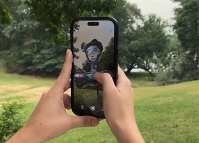 Hands holding a smartphone outdoors showing an augmented reality sculpture of a colorful human-like bust on the phone screen, set against a grassy park with trees.