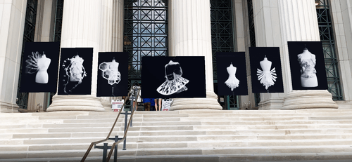 Art exhibition display outside a building with large stone columns, featuring seven augmented reality framed photos of avant-garde white geometric and abstract dress designs on mannequins, arranged above stone steps.